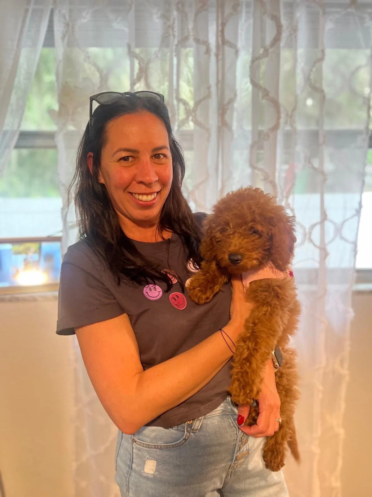 A woman with dark hair, glasses on her head, smiling, holding a small brown poodle puppy with curly fur, inside a room with sheer curtains and windows in the background.