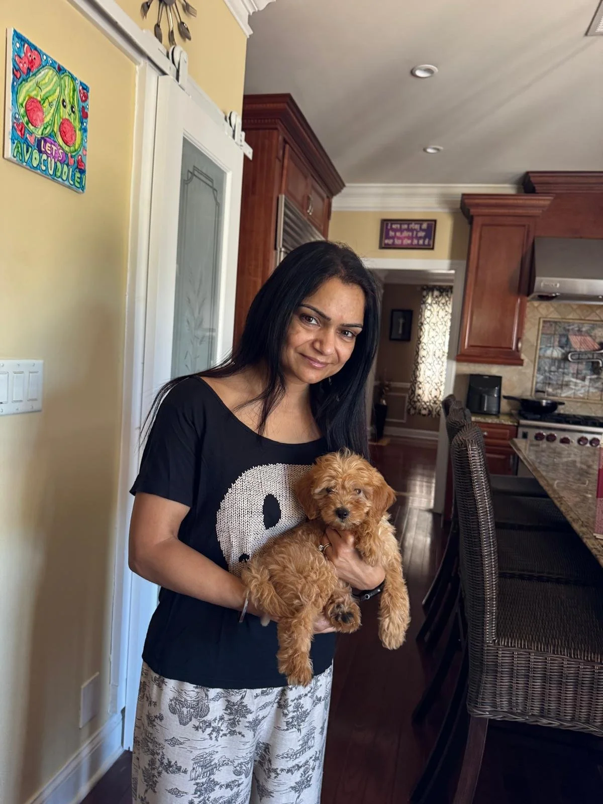A woman with long black hair wearing a black t-shirt featuring a white skull design is holding a small, fluffy, light brown puppy inside a kitchen.