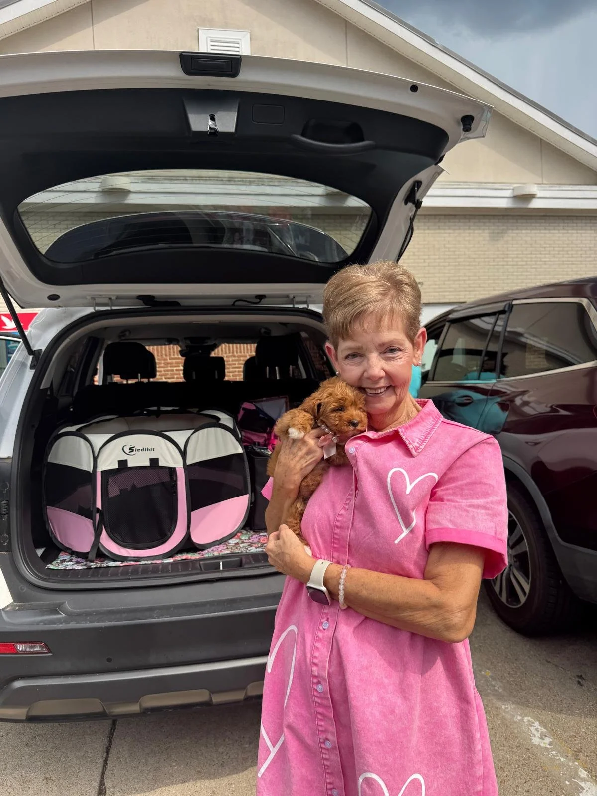 A smiling woman holding a small brown puppy in front of an open car trunk filled with pet carriers and supplies, parked in a driveway with houses in the background.