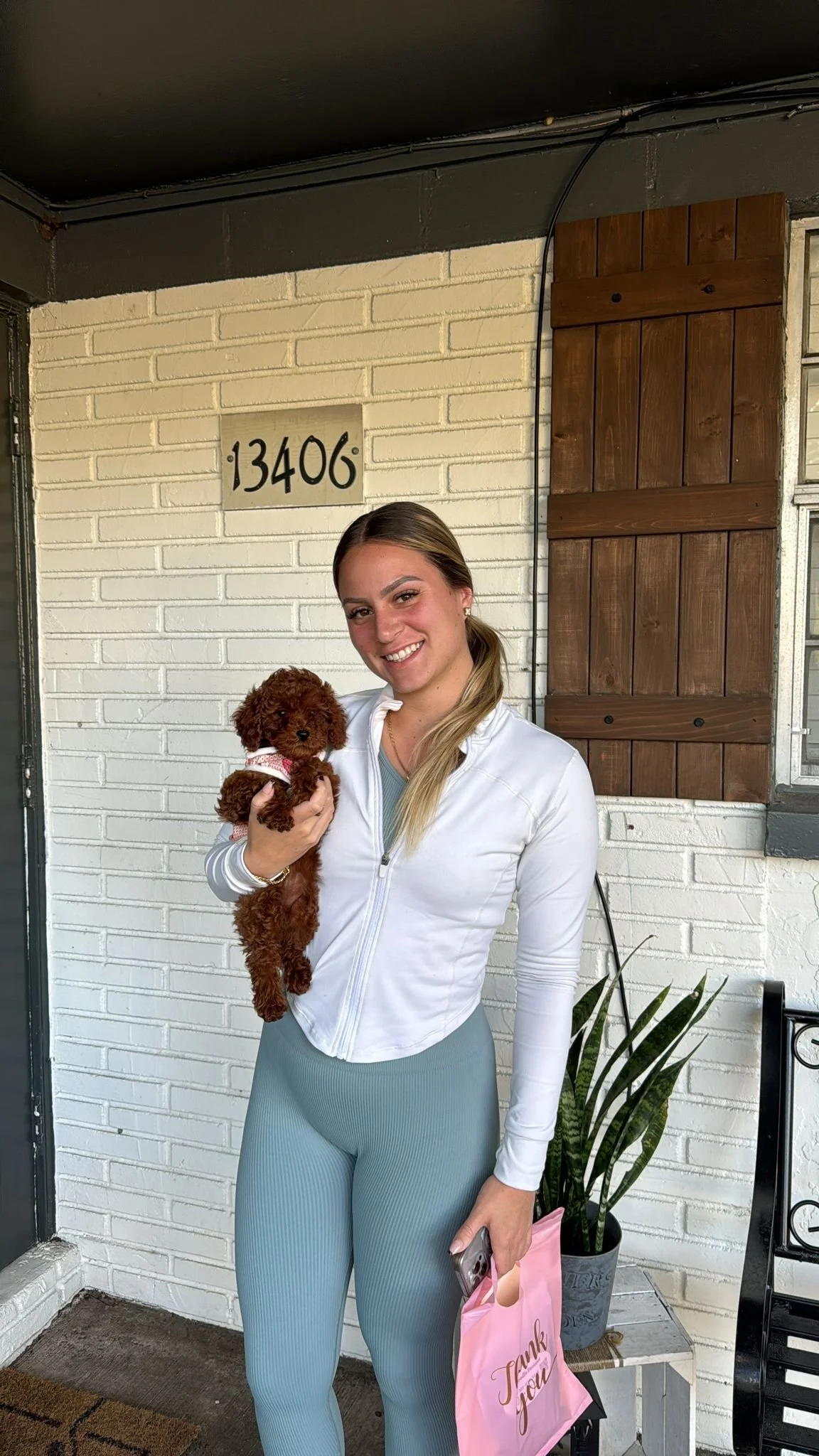A woman with long blonde hair holding a small curly-haired brown puppy with a bowtie, standing in front of a white brick wall with the address 13406. She is smiling, wearing a white zip-up jacket and light blue leggings, and is holding a pink gift ba