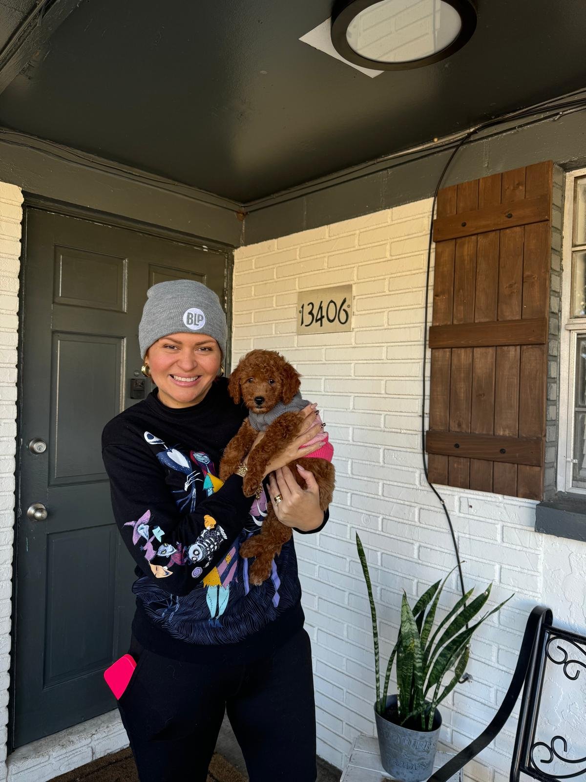 A woman smiling and holding a small brown puppy on a porch in front of a white brick wall with the house number 13406. The woman is wearing a gray beanie and a black sweater with colorful designs. There is a potted snake plant and a metal chair with 