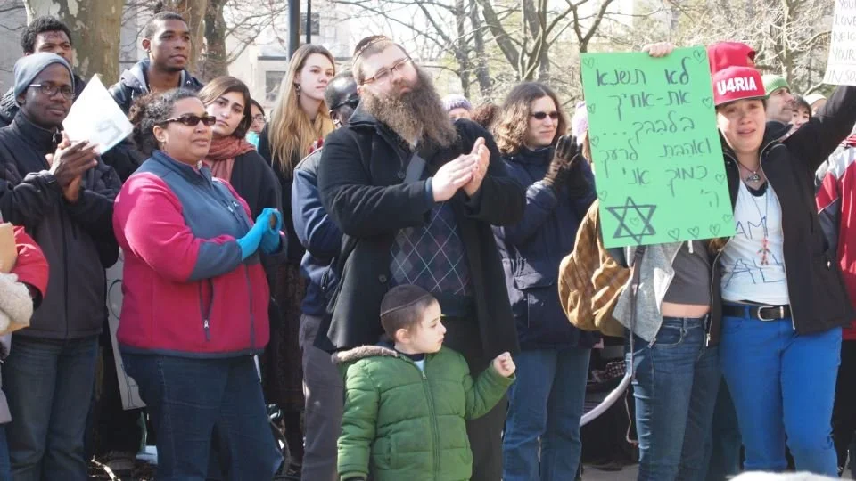 A diverse group of people gathered  with Shlomo Elkan outdoors at a protest or rally, with some holding signs and others clapping or observing.
