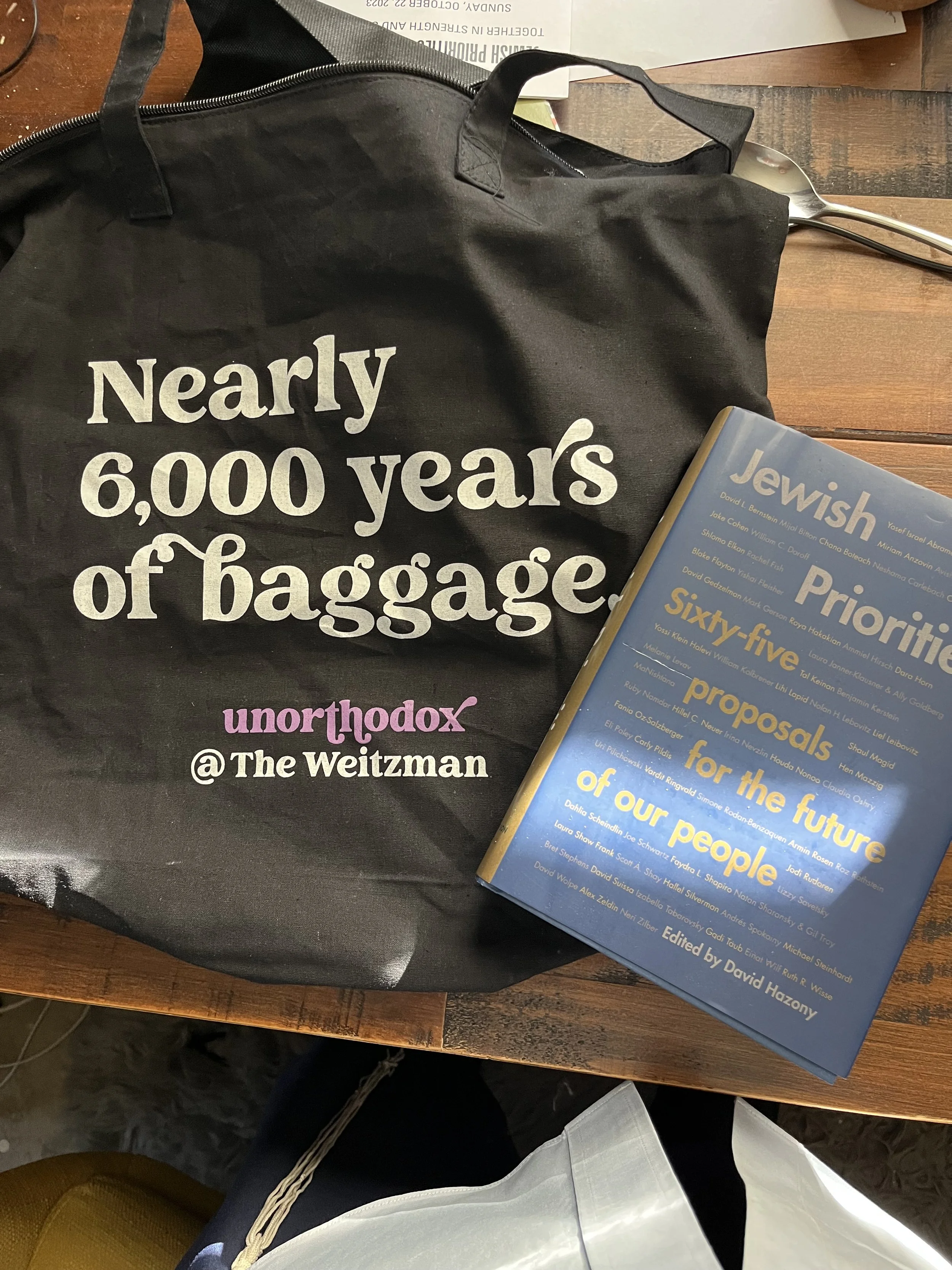 A black tote bag with white text that reads 'Nearly 6,000 years of 'baggage' unorthodox @The Weitzman', a blue book titled 'Jewish Priorties' is placed on a wooden table alongside a silver spoon and some papers.