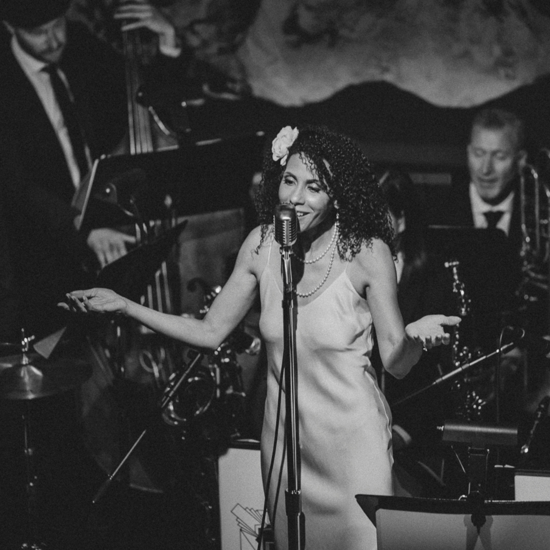 A woman with curly hair, wearing a flower hair accessory, pearl necklace, and sleeveless dress, singing into a vintage microphone on stage with jazz band musicians in the background.