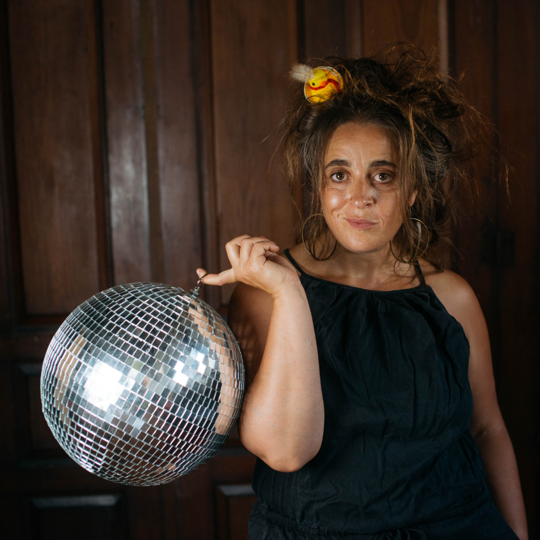 A woman with messy hair holding a disco ball in her right hand, standing against a wooden-paneled wall, wearing a black sleeveless top, with a small plush yellow rubber duck on her head.