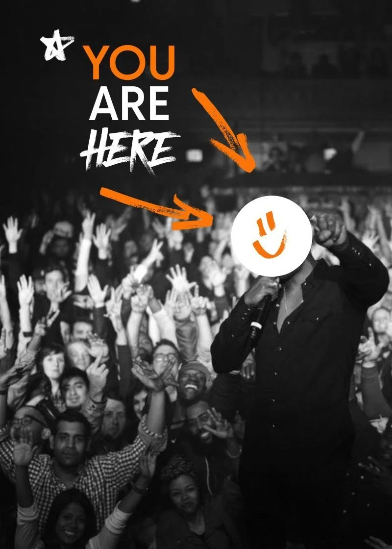 Black and white photo of a large crowd of people at a concert or event, raising their hands, with a person in the foreground holding a smiley face mask in front of their face. The image has colorful text that says 'You Are Here' with arrows pointing to the person's face.