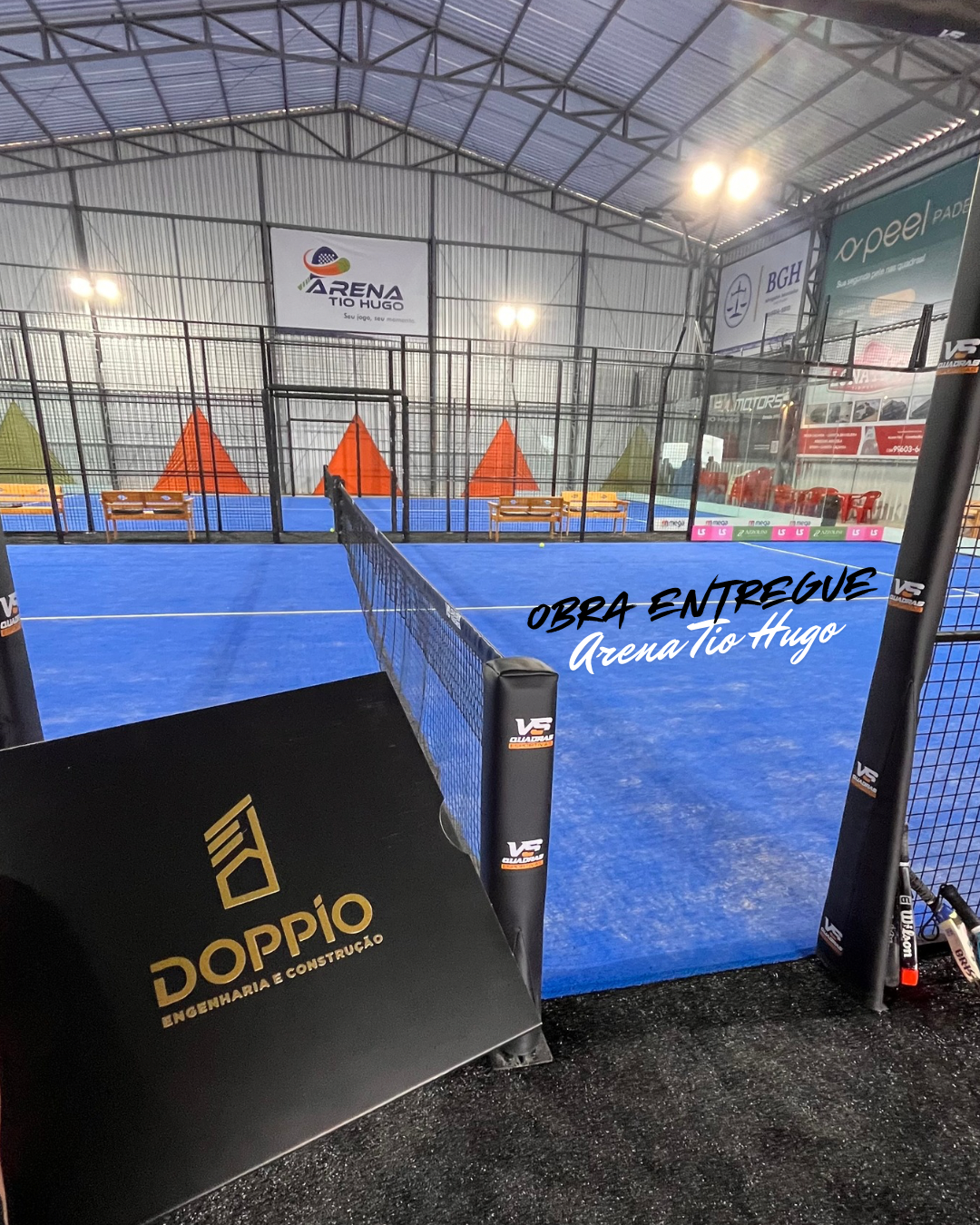 Quadra de beach tennis com piso azul, cercada por uma grade de metal preta, com alguns bancos de madeira ao fundo e cones de sinalização de cores diferentes ao lado. Na parede ao fundo, um painel com o nome 'Arena Tio Hugo' e logos de patrocinadores,