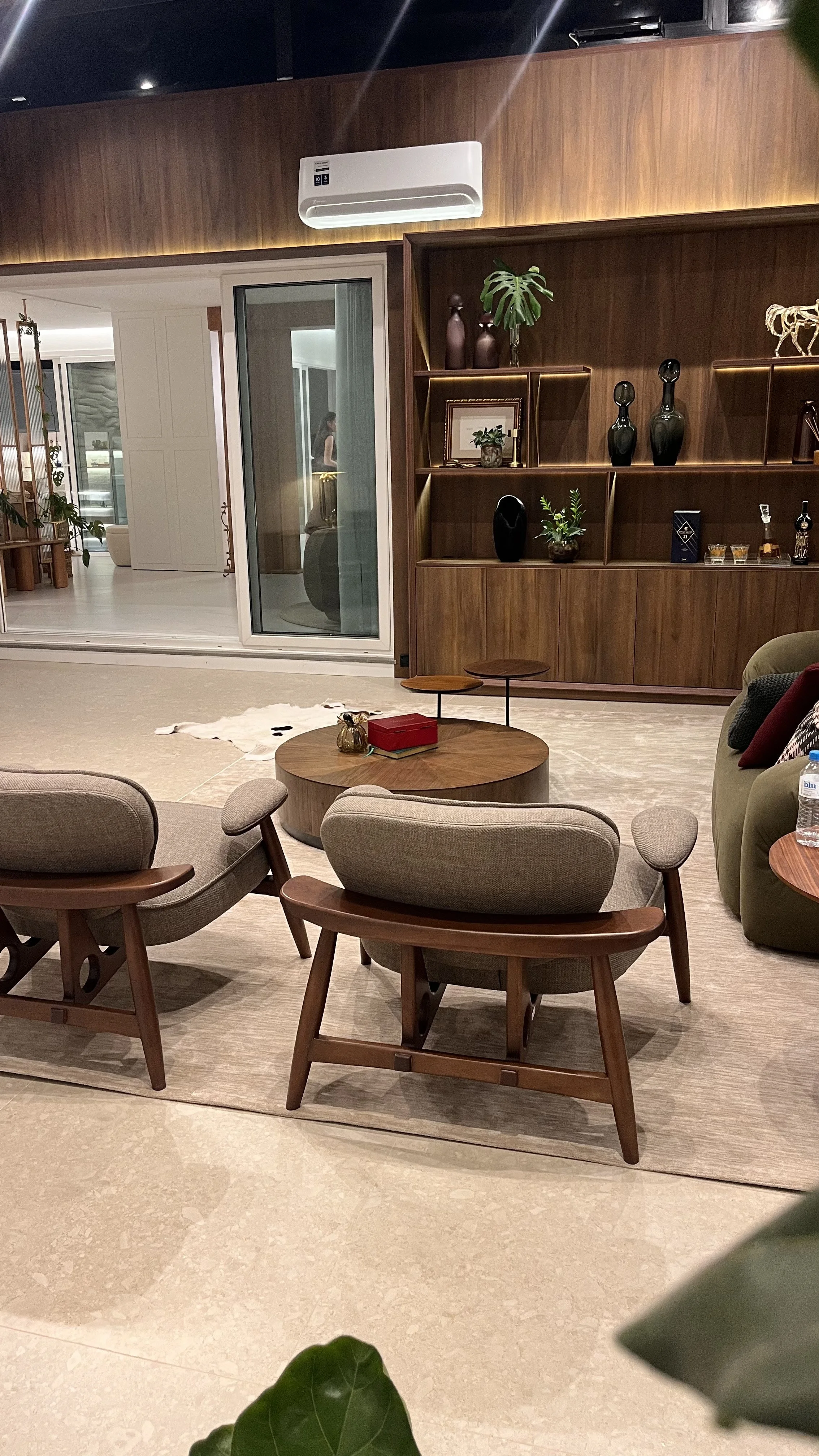 Sala de estar decorada com móveis de madeira, incluindo uma estante com vasos decorativos e plantas. Há uma mesa de centro de madeira com objetos decorativos e uma poltrona. Ao fundo, uma divisória de vidro separa a sala de um espaço com mais móveis 