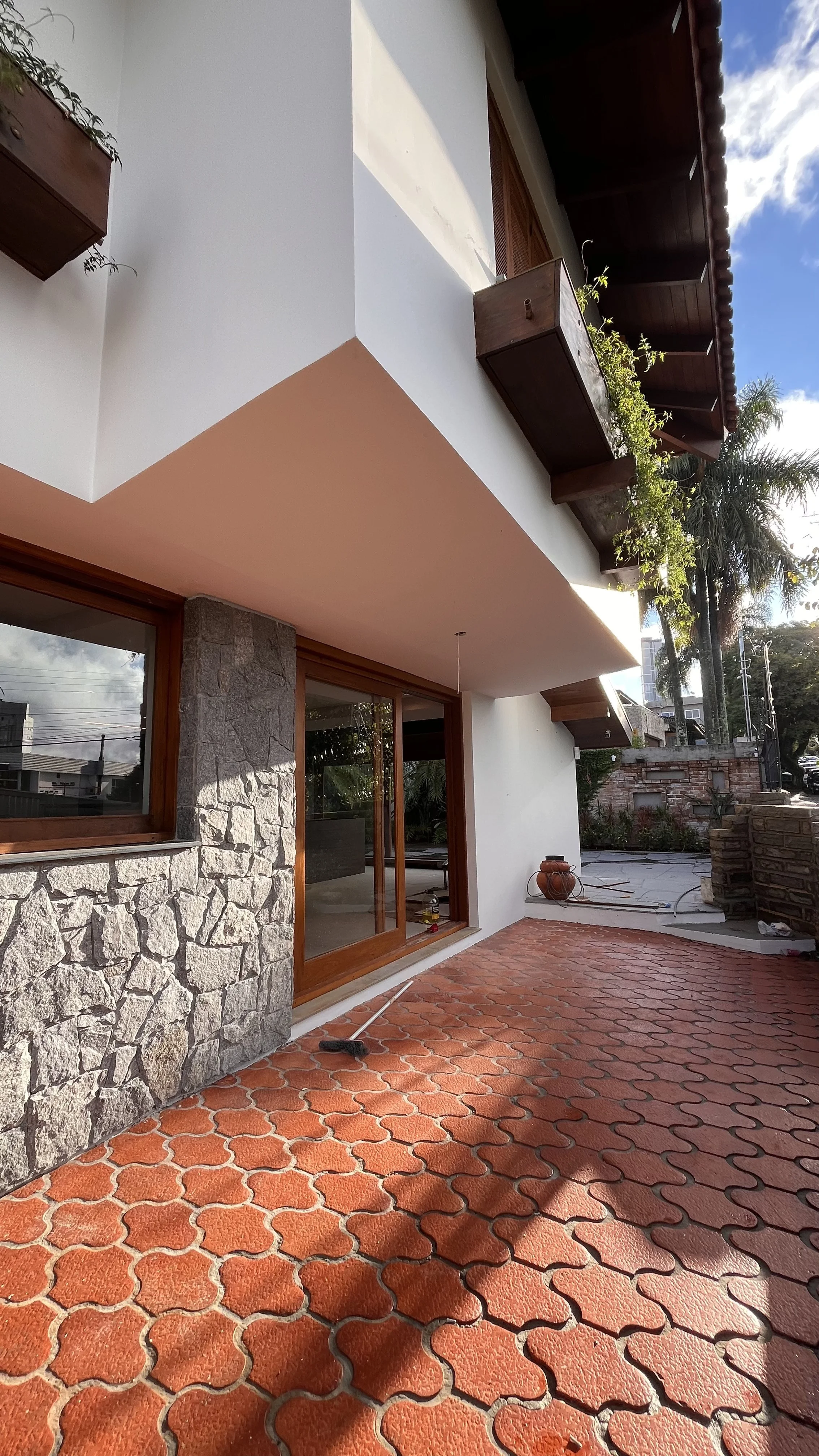 Fachada de uma casa com piso de ladrilho vermelho, parede branca, janela de vidro com moldura de madeira, parede de pedra e varanda com vaso de plantas e ferramenta de limpeza