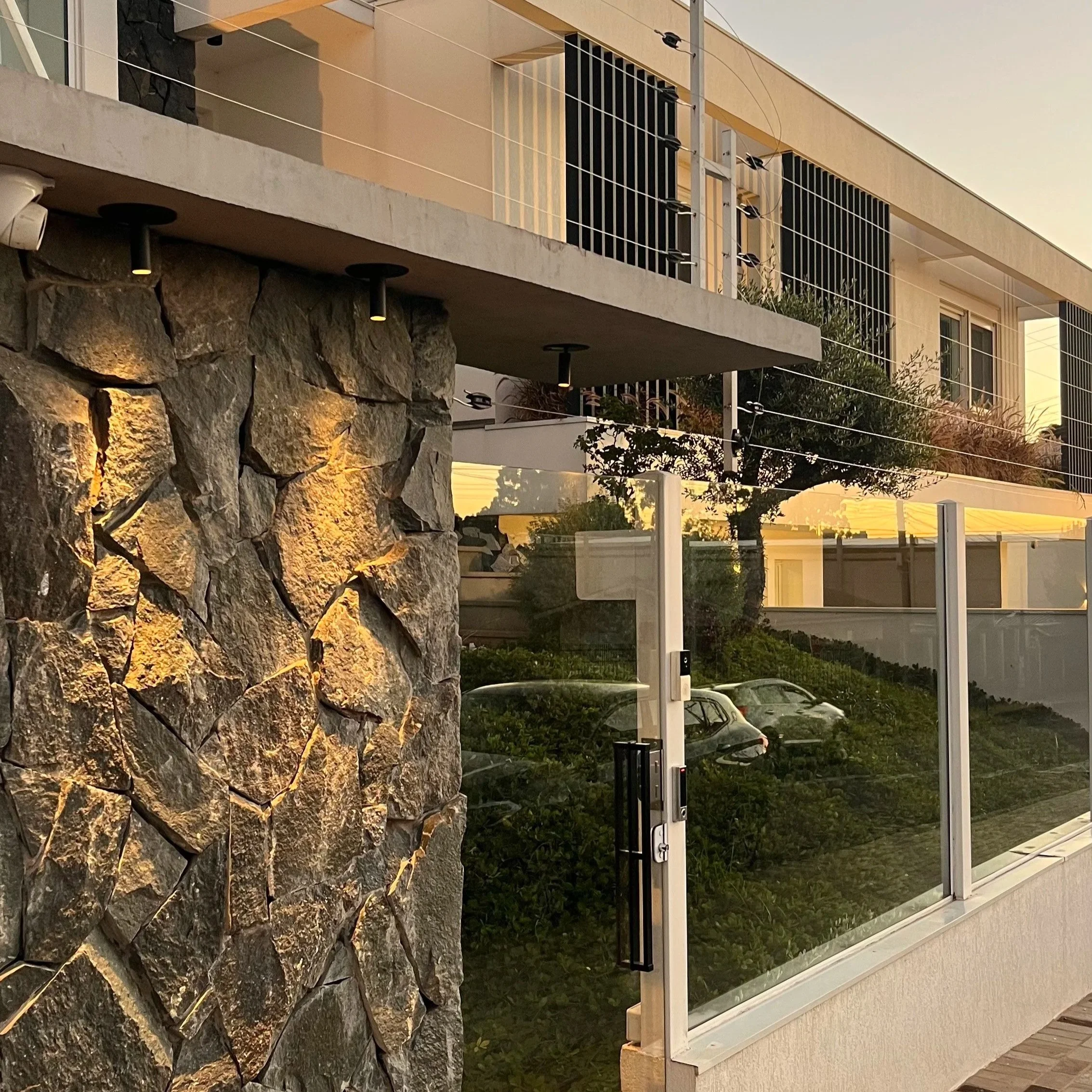 Fachada de uma casa moderna com muro de pedras iluminado, janelas com grades pretas, varanda com vidro, árvores e céu ao entardecer.