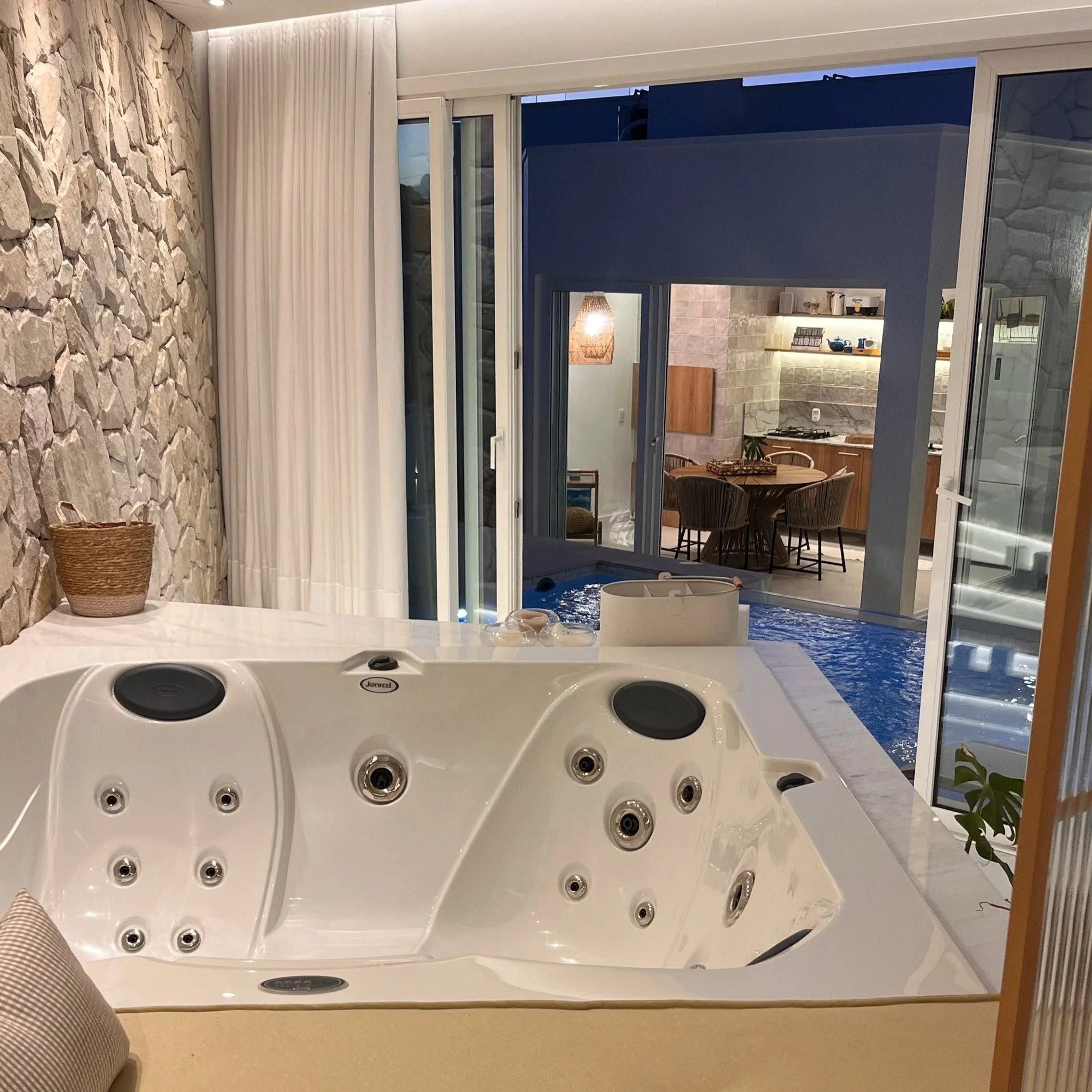 Banheira de hidromassagem branca com múltiplos jets, em um ambiente interno, com uma parede de pedra na esquerda e uma parede azul ao fundo, vista de cima.