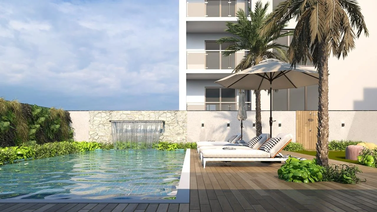 Piscina com espelho d'água e cascata, cadeiras de descanso com guarda-sol e plantas tropicais ao redor, ao fundo um prédio residencial.
