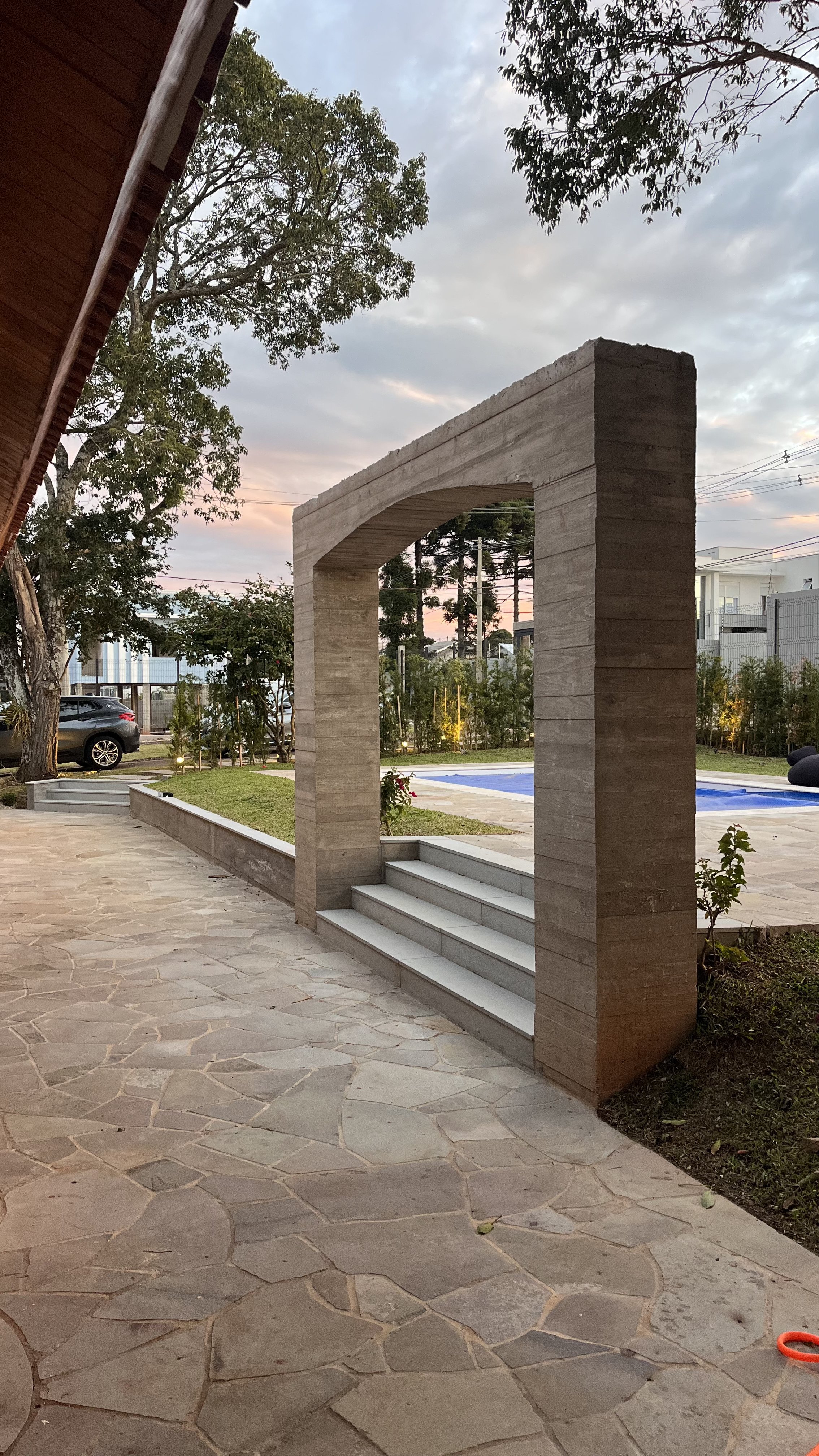 Estrutura de concreto com escadas, jardim com plantas, piscina ao fundo, árvores altas e céu com nuvens ao entardecer.