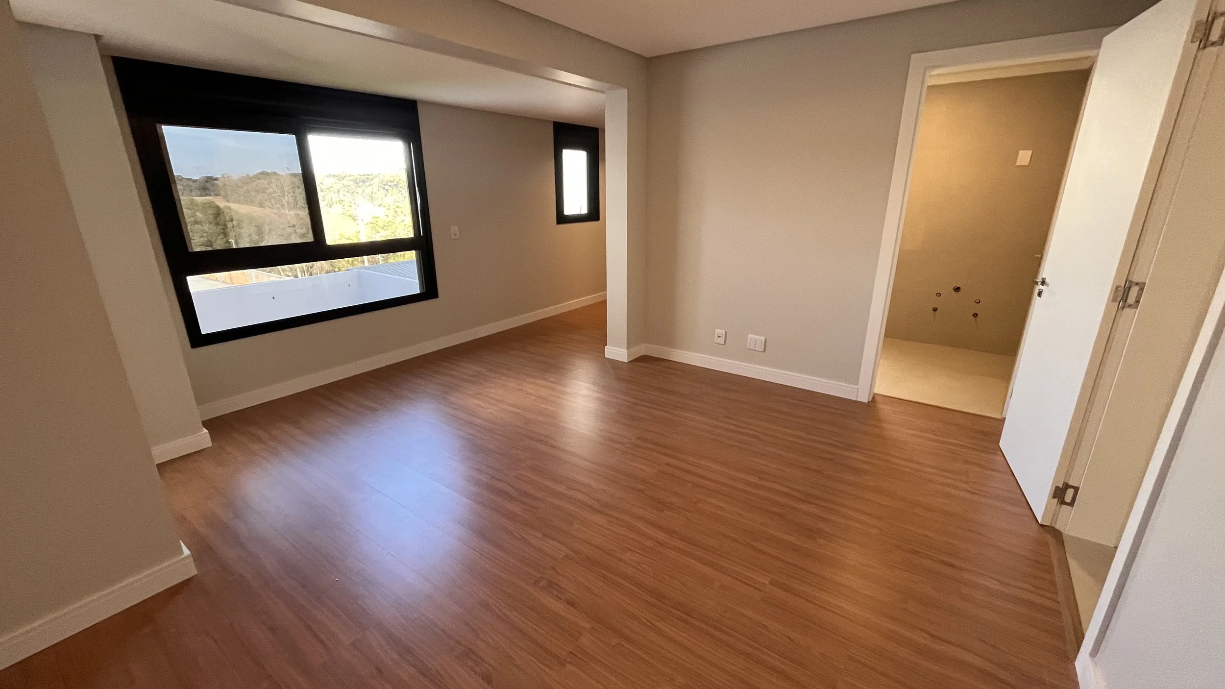 Sala vazia com piso de madeira, parede cinza-claro, janela grande de vidro com moldura preta e uma porta aberta que revela uma área de serviço ou banheiro com parede amarela pálida.