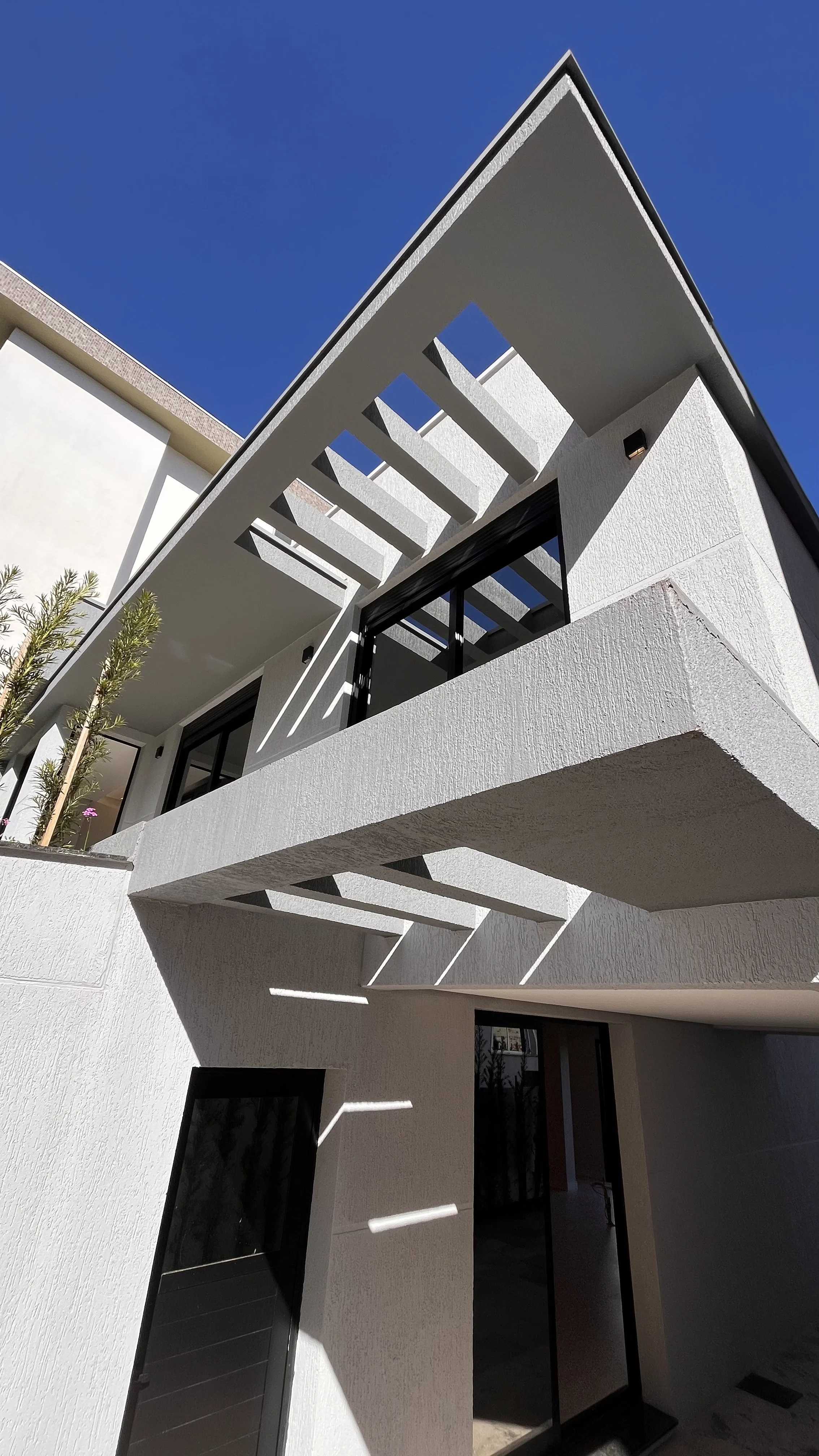Fachada de uma casa moderna com varanda suspensa e detalhes em branco e preto, sob céu azul claro.