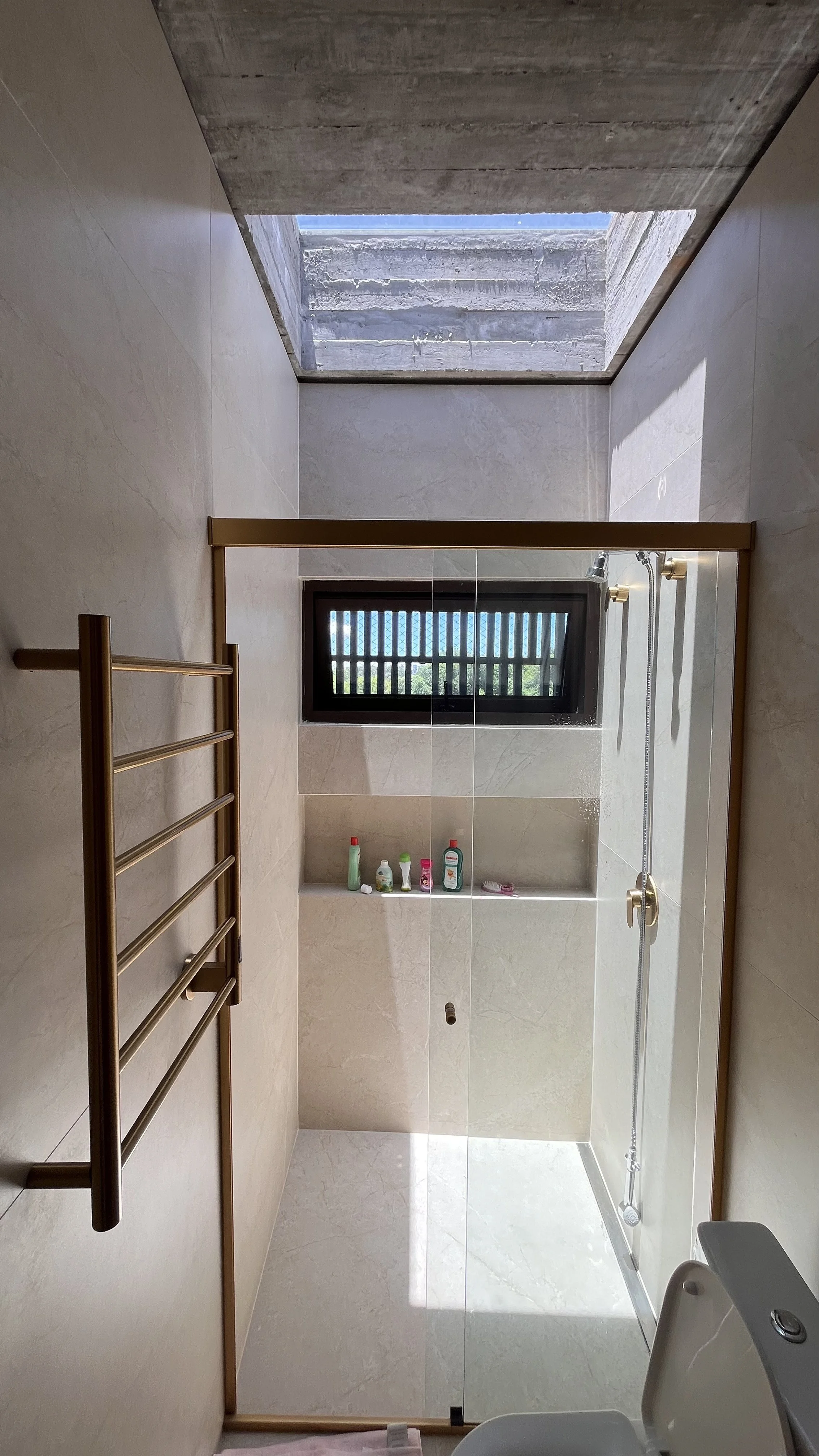 Banheiro com parede de azulejo claro, janela de vidro com grades, ducha com suporte e produtos de banho na bancada. Persiana na janela e teto de concreto com clarabóia aberta.