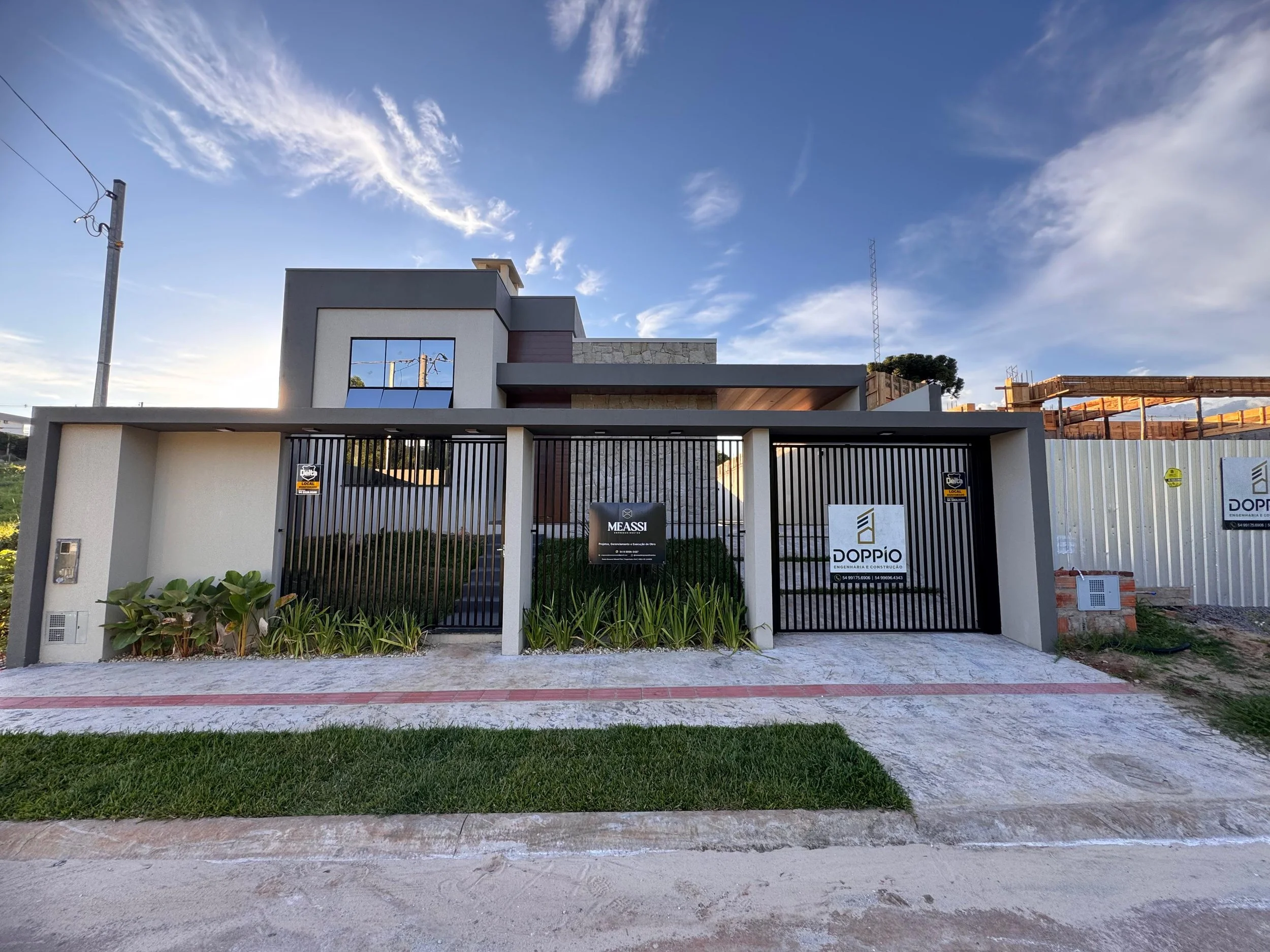 Fachada de uma casa moderna com detalhes em pedra, bancada de vidro com cercas de ferro preto na frente, jardim com plantas verdes e sinalização de construção no lado direito, céu com nuvens brancas e sol ao fundo.