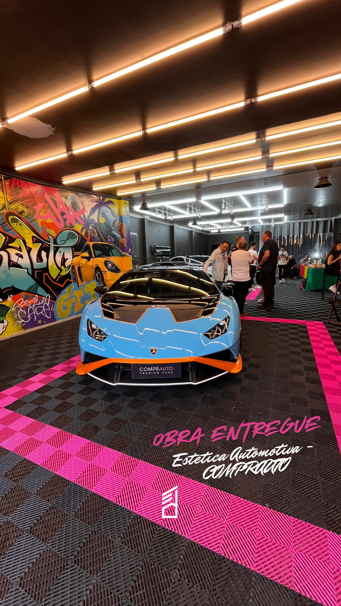Carro esportivo azul com detalhes laranja em um evento de automóveis, com pintura de graffiti ao fundo e pessoas ao redor observando, piso com marcações rosa, ambiente com iluminação moderna.