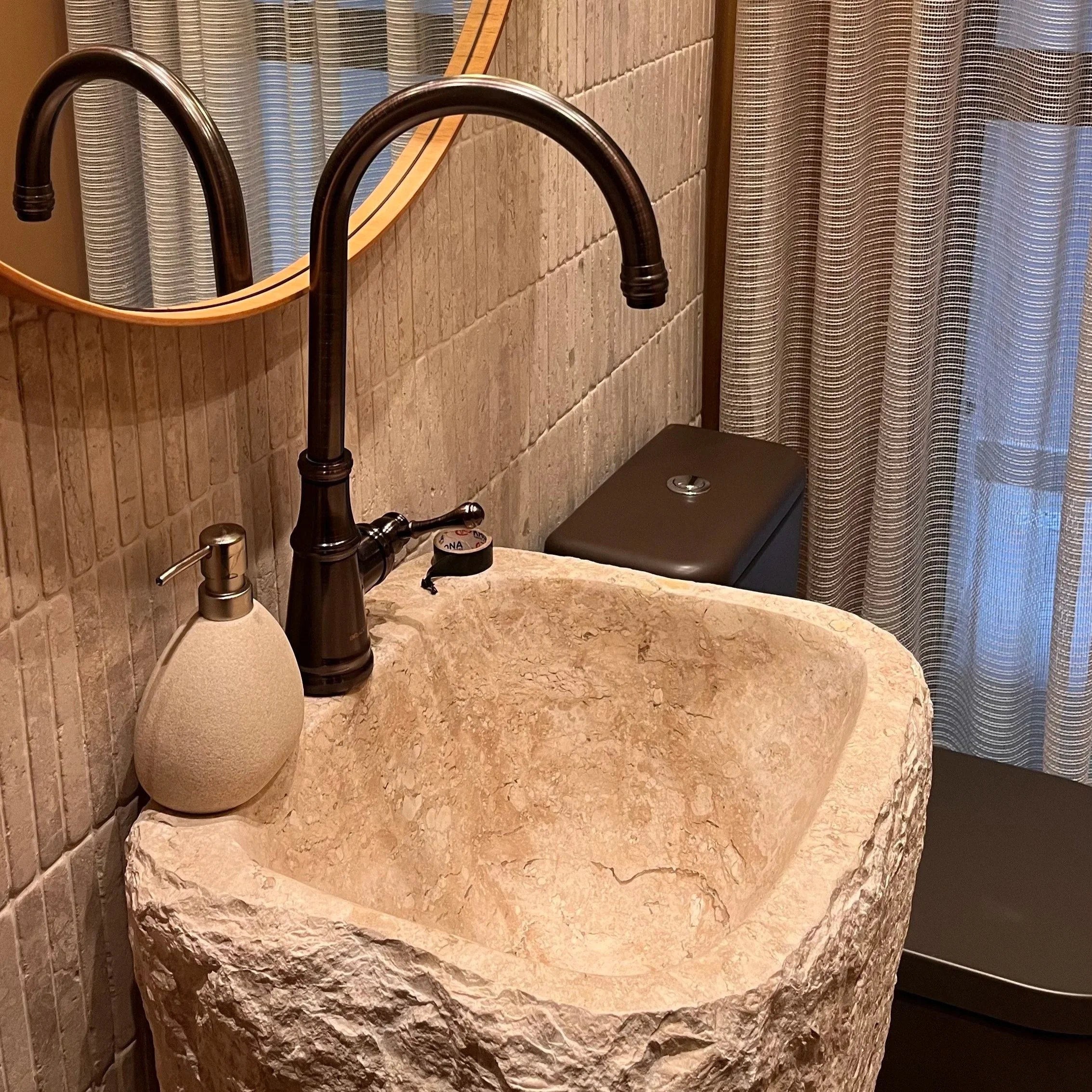 Imagem de um banheiro com pia de pedra, espelho, torneira preta, cortina de banheiro listrada e um vaso sanitário ao fundo.