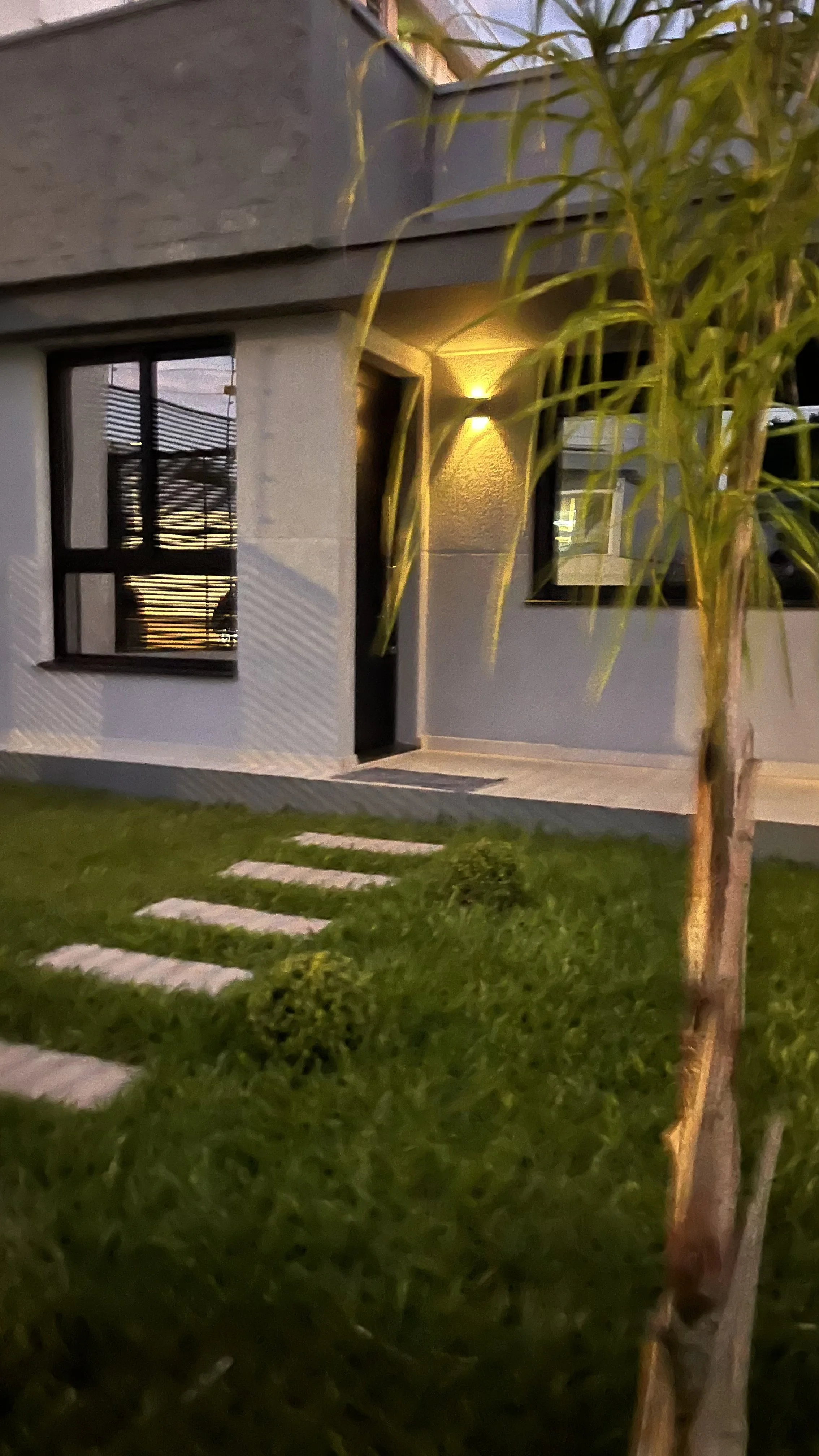 Imagem de uma casa moderna vista de fora ao entardecer, com iluminação acesa na parede, uma janela grande e uma porta de entrada, tendo um jardim com grama e um caminho de pedras na frente, e uma árvore ao lado esquerdo.