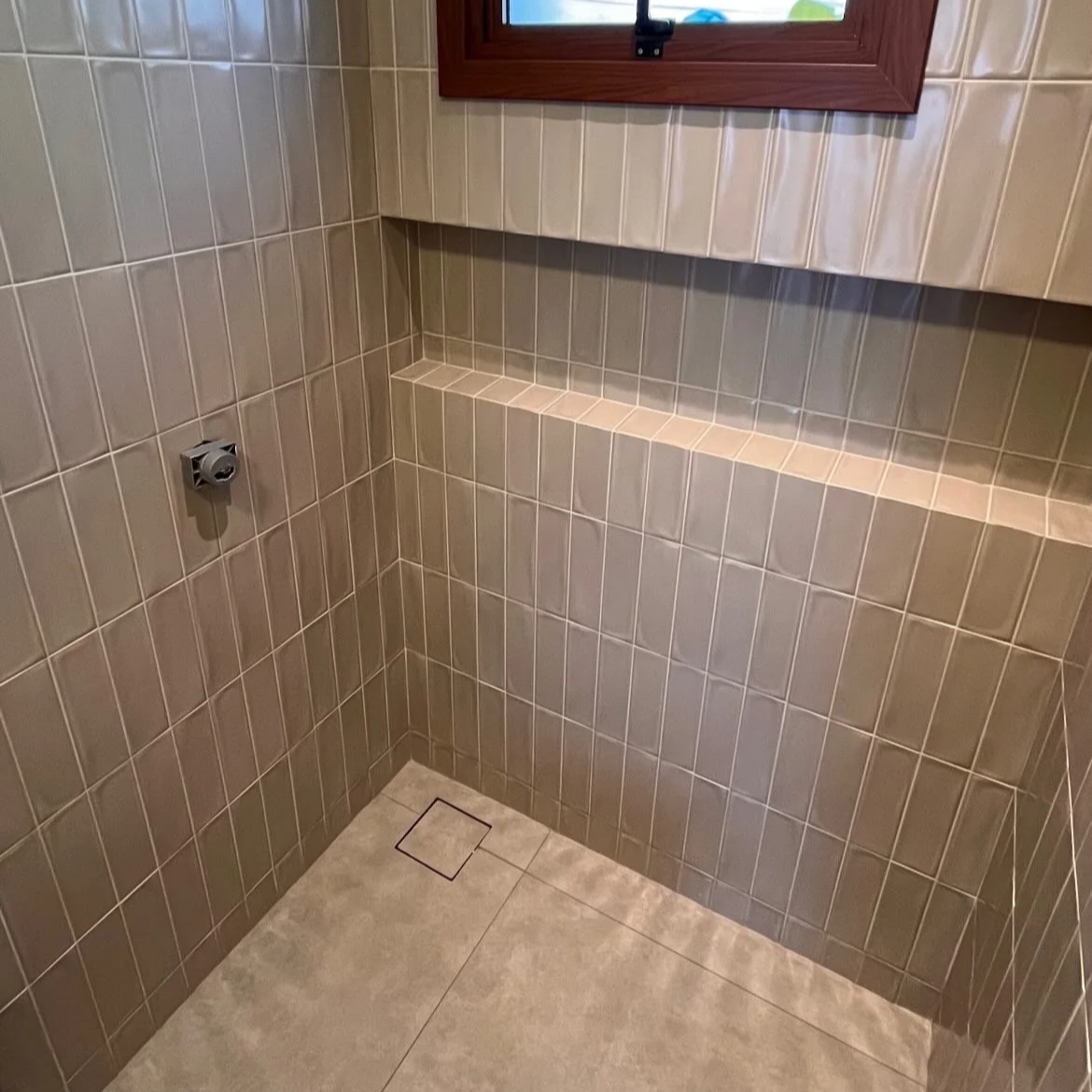 Área de banho com paredes revestidas de azulejos bege e piso de cerâmica clara. Há uma pequena prateleira embutida na parede e uma janela de vidro com moldura de madeira.