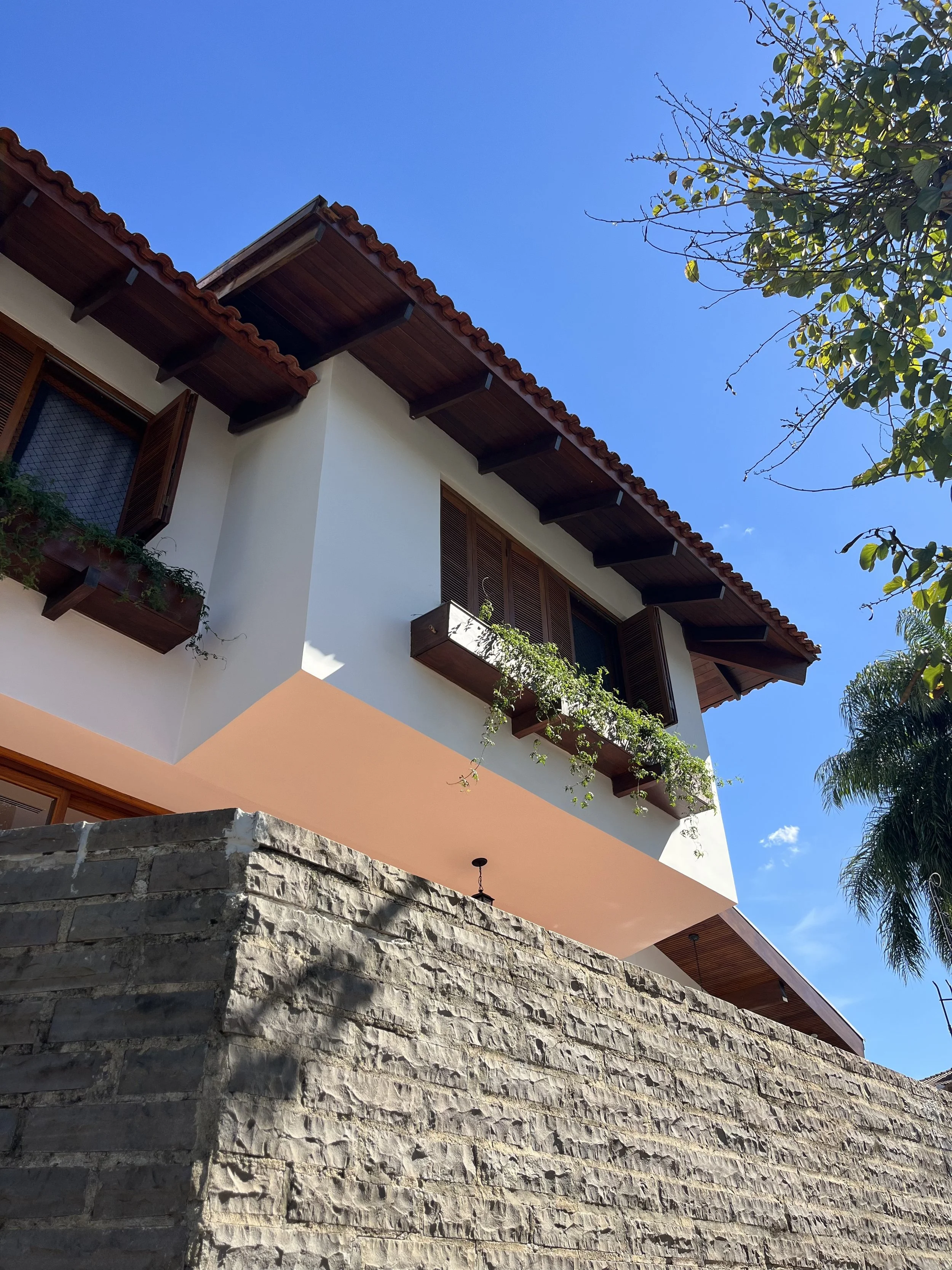 Fachada de uma casa moderna com parede de pedra, janelas de madeira com persianas, e um terraço com plantas, sob céu azul claro com algumas árvores ao redor.