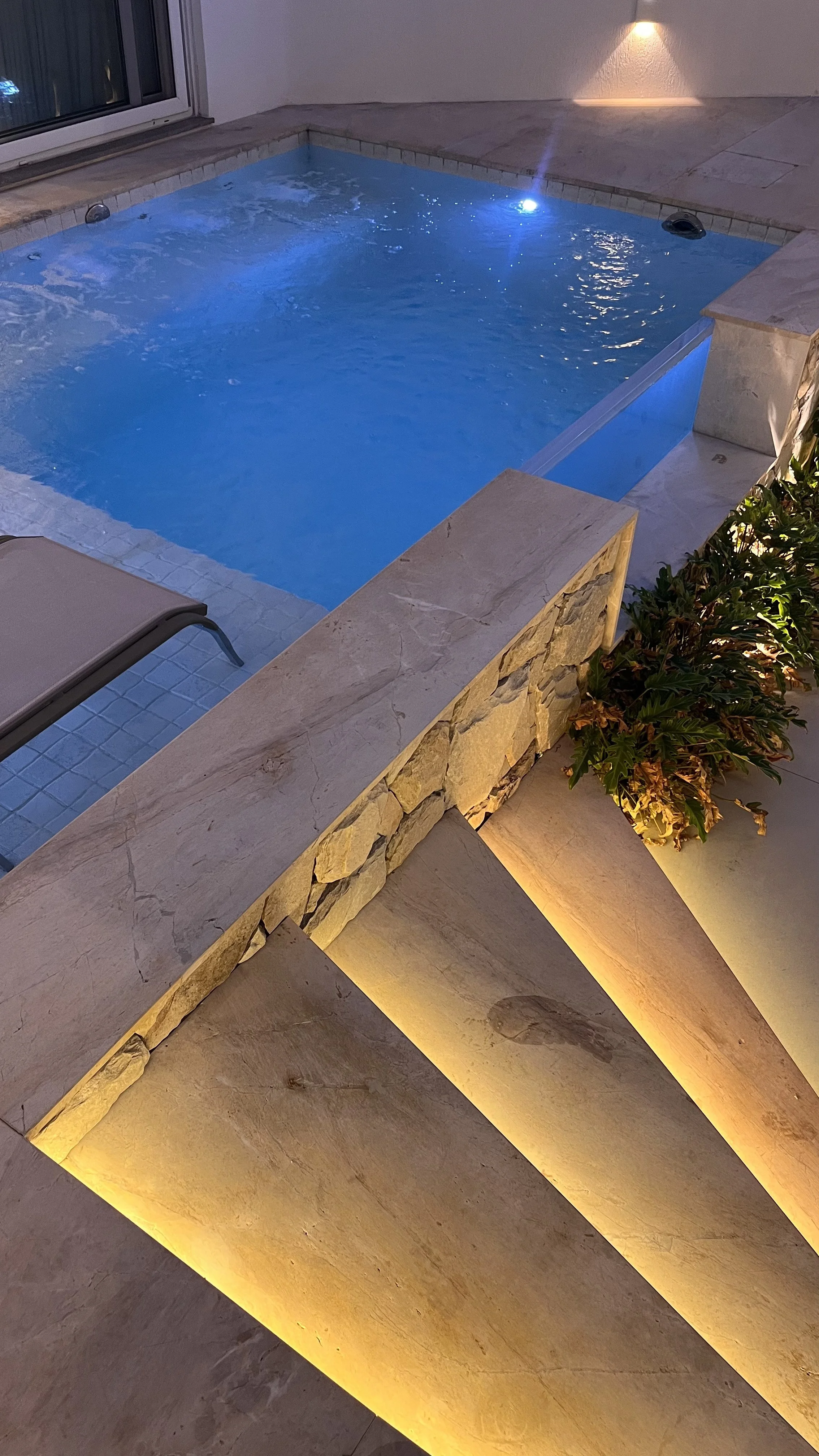 Piscina moderna com iluminação noturna, cercada por uma borda de pedra e com plantas decorativas ao lado.