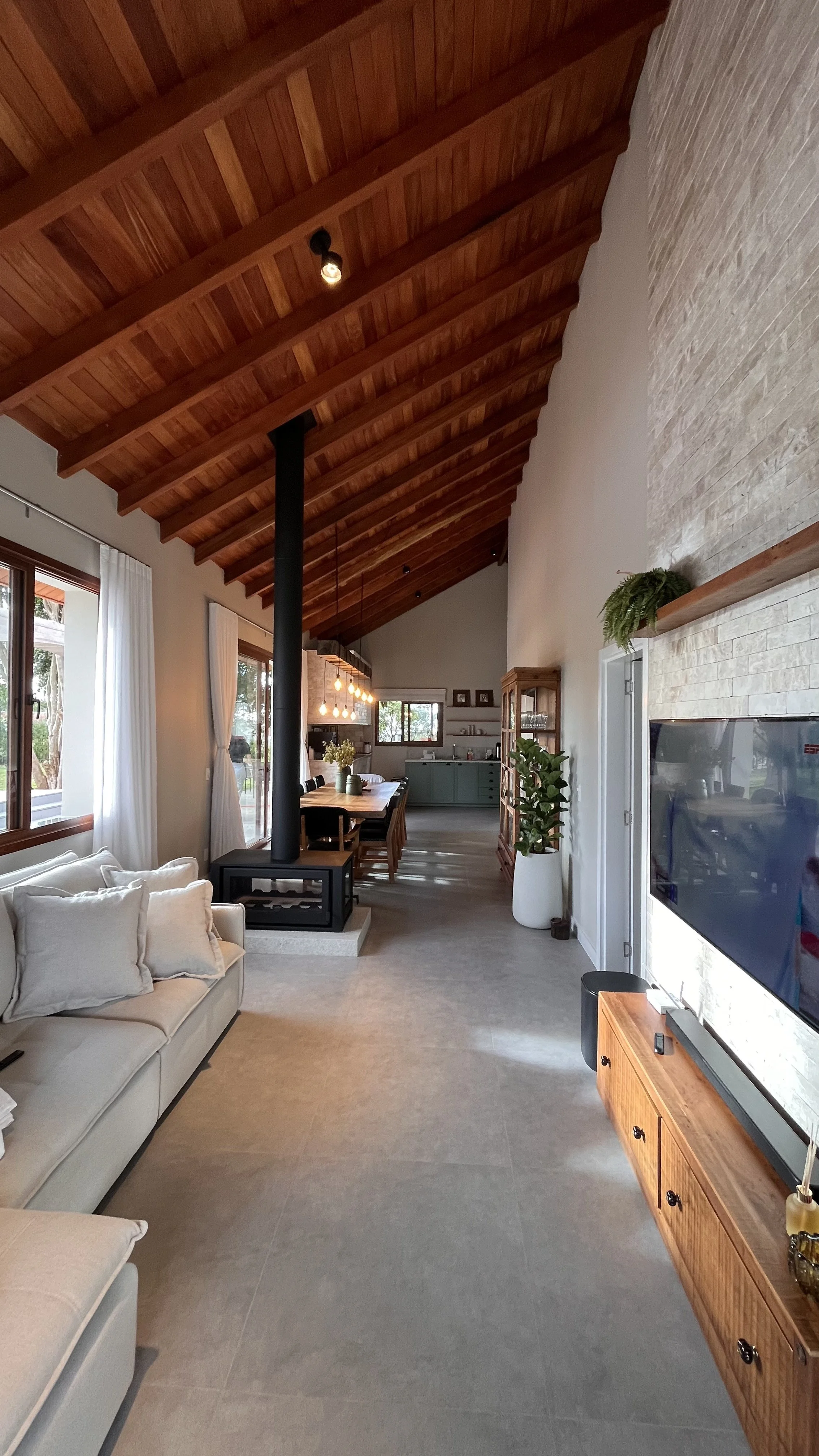 Sala de estar com teto arqueado de madeira, parede de pedra , sofá bege com almofadas, janelas grandes com cortinas brancas, área de jantar com mesa e cadeiras, cozinha ao fundo com armários azuis, plantas decorativas, televisão na parede e piso de ladrilhos cinza.