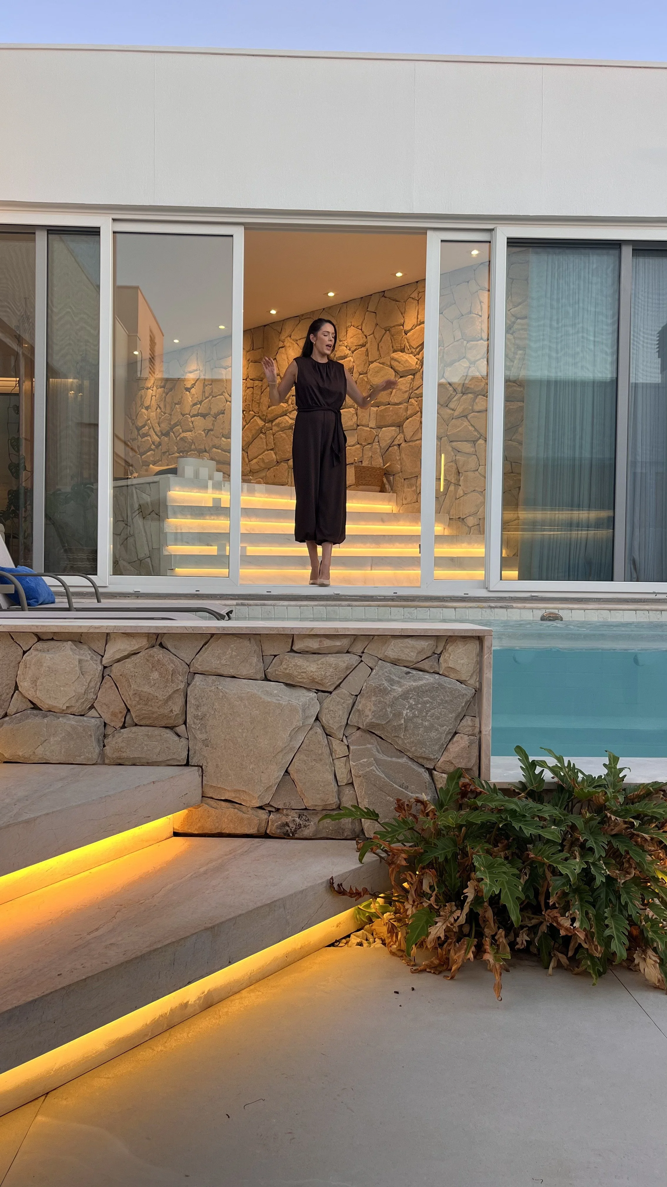 Mulher de vestido preto de pé dentro de uma casa de vidro com paredes de pedra, ao fundo escadas com iluminação amarela e uma piscina ao lado.
