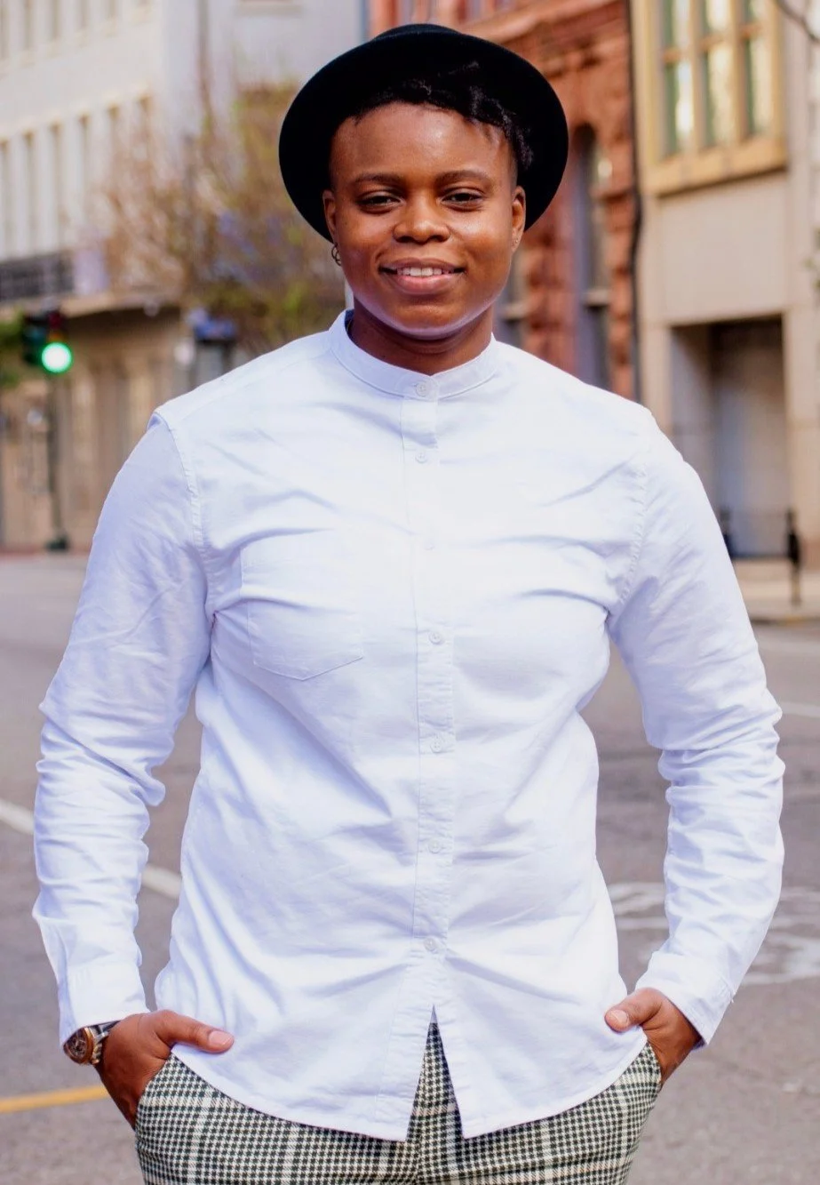 A person standing outdoors on a city street, wearing a white button-up shirt, patterned pants, a black hat, and a wristwatch, smiling with hands in pockets.