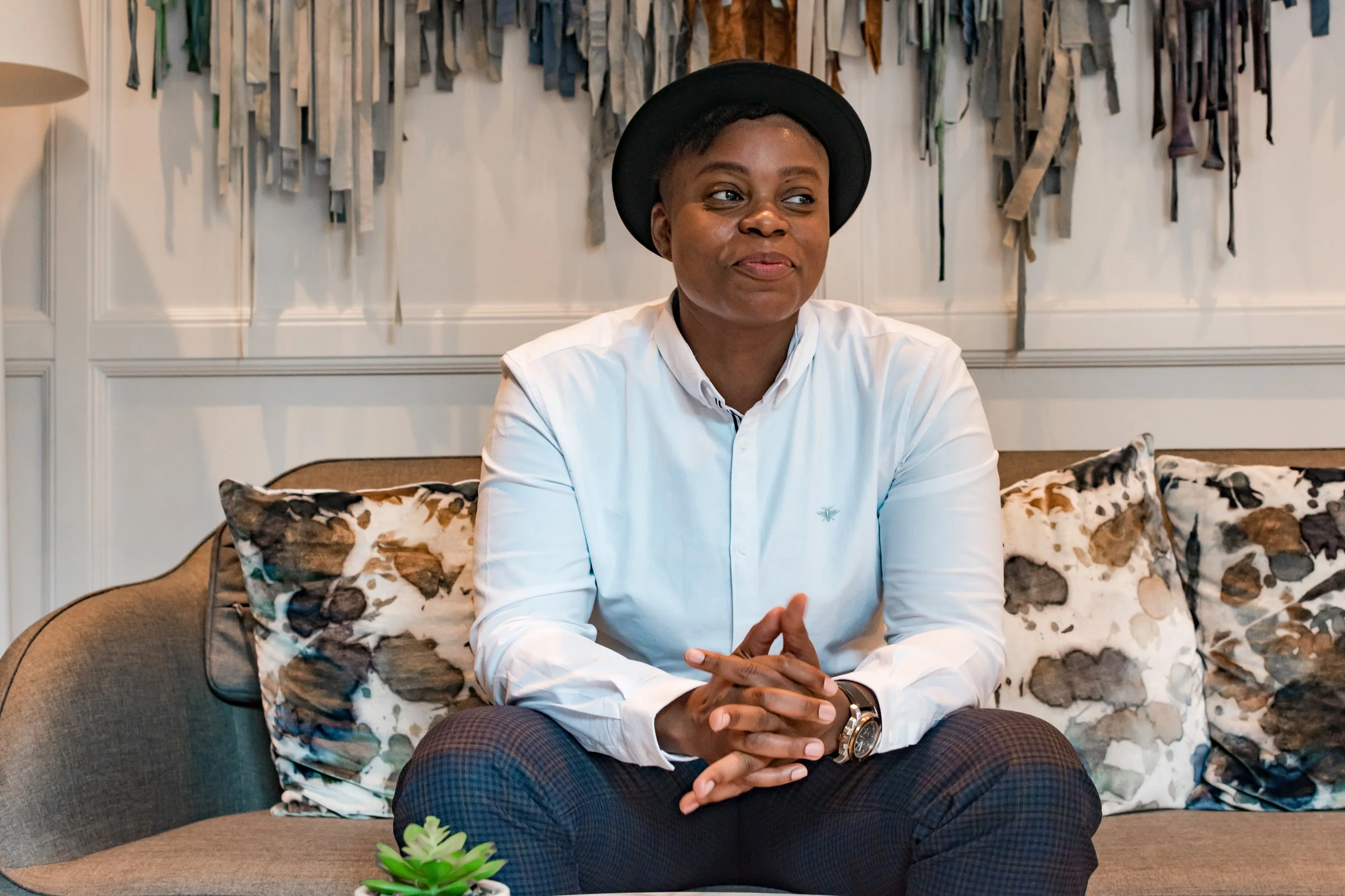 A woman sitting on a sofa with patterned throw pillows, wearing a white shirt and a black hat, smiling slightly with hands clasped, in a room with white panel walls and a colorful textile wall hanging.