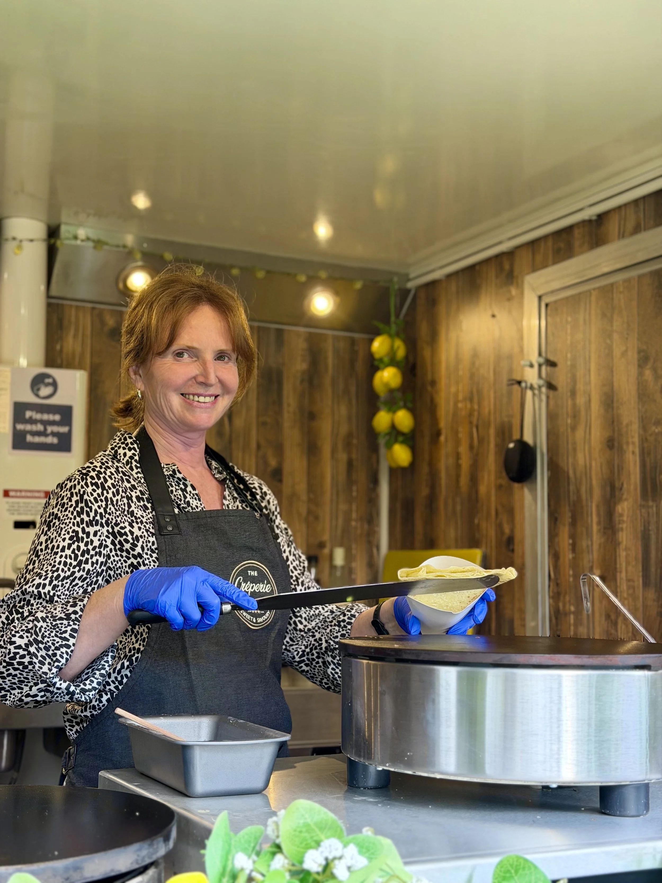 A woman with short red hair and a leopard print shirt smiling while serving crepes at a food stall. She is wearing a black apron and blue gloves, holding a spatula and a white dish. The background has wooden walls, yellow lemons hanging, and warm lighting.