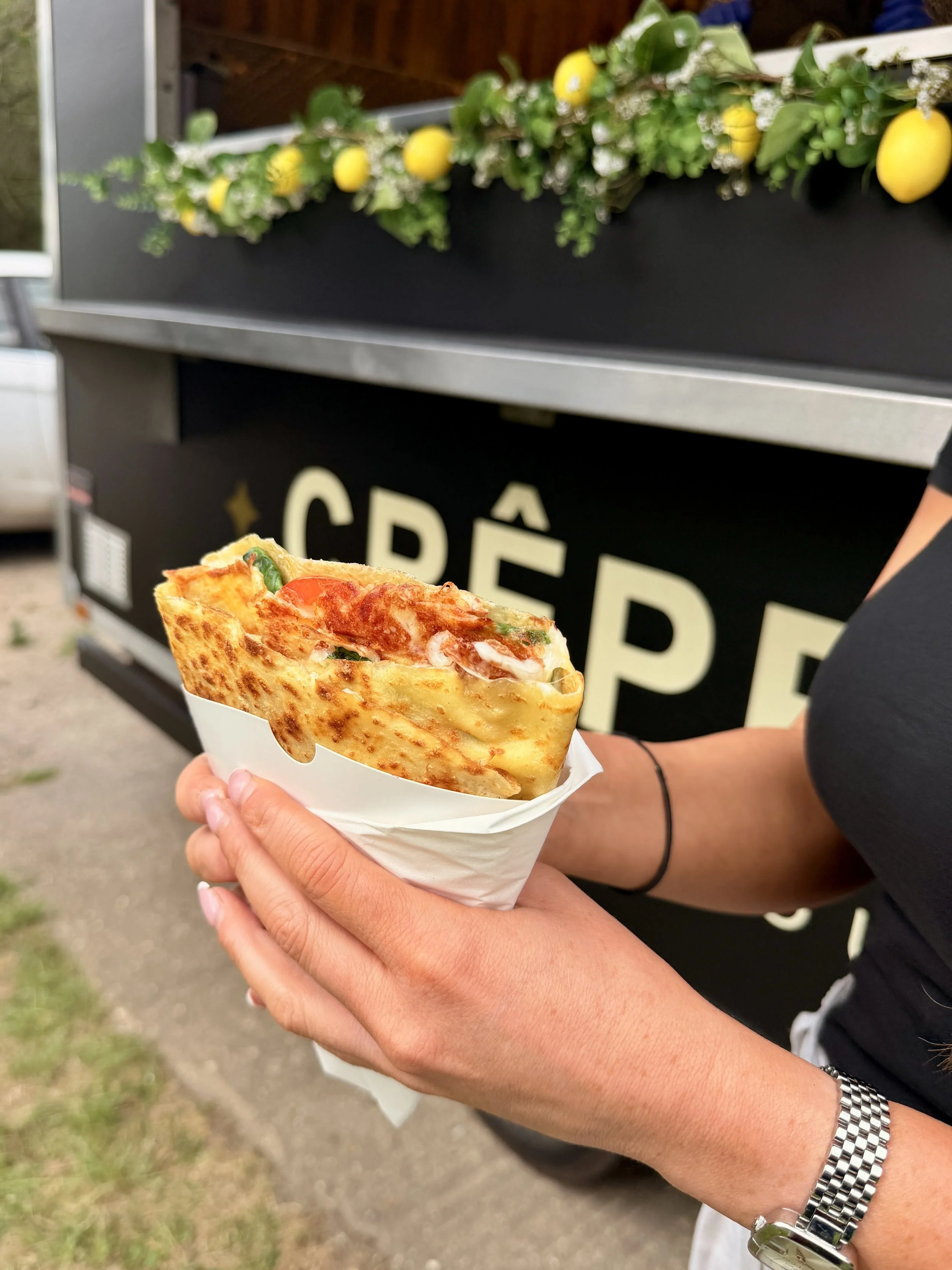 A person holding a crepe with visible fillings including cheese, tomatoes, and ham, in front of a food stand decorated with yellow lemons and white flowers.