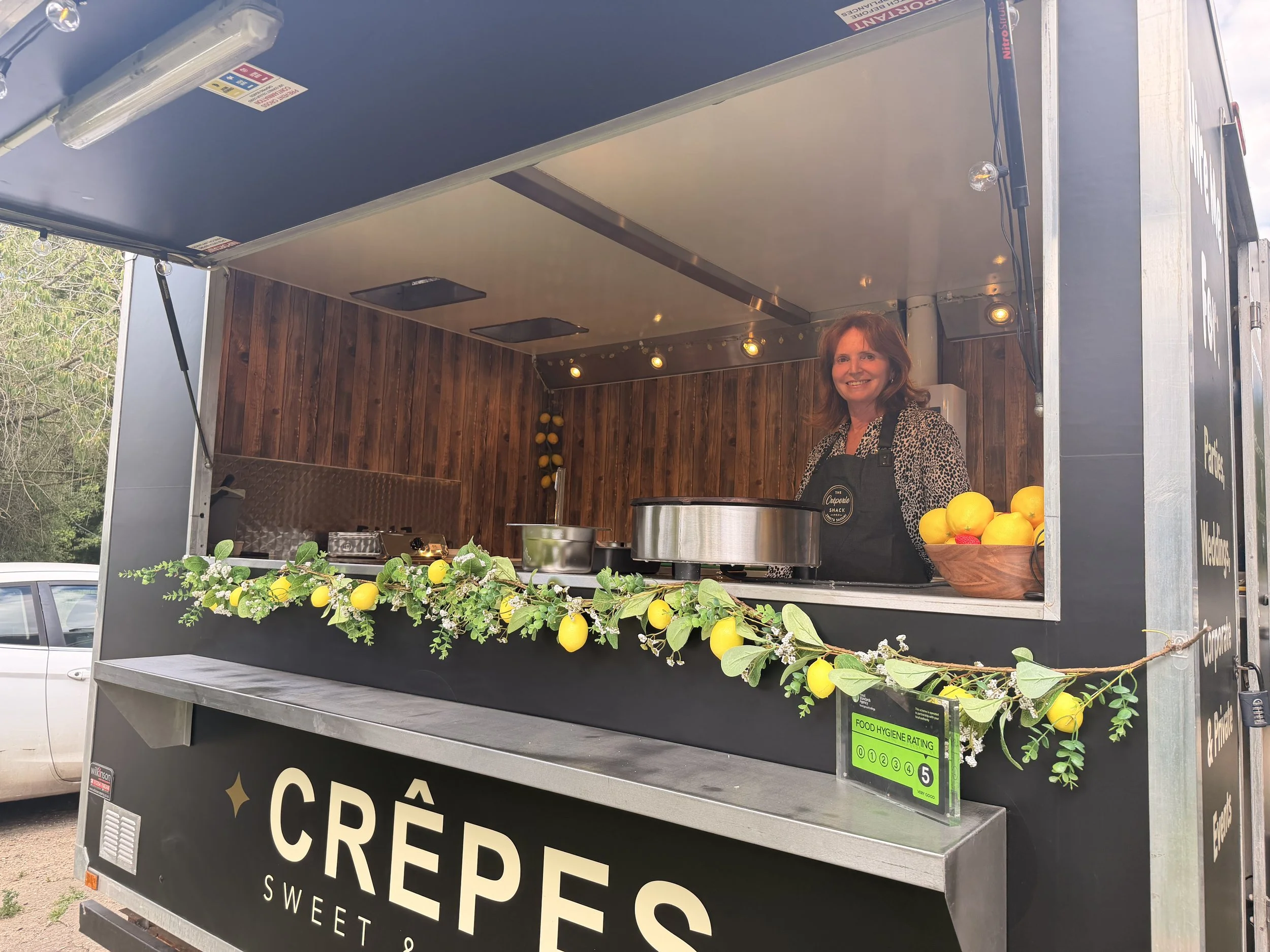 A woman standing inside a food truck with wooden paneling and a black sign that reads 'Crêpe'. The food truck is decorated with lemons and greenery, and the woman is smiling and wearing a black apron.