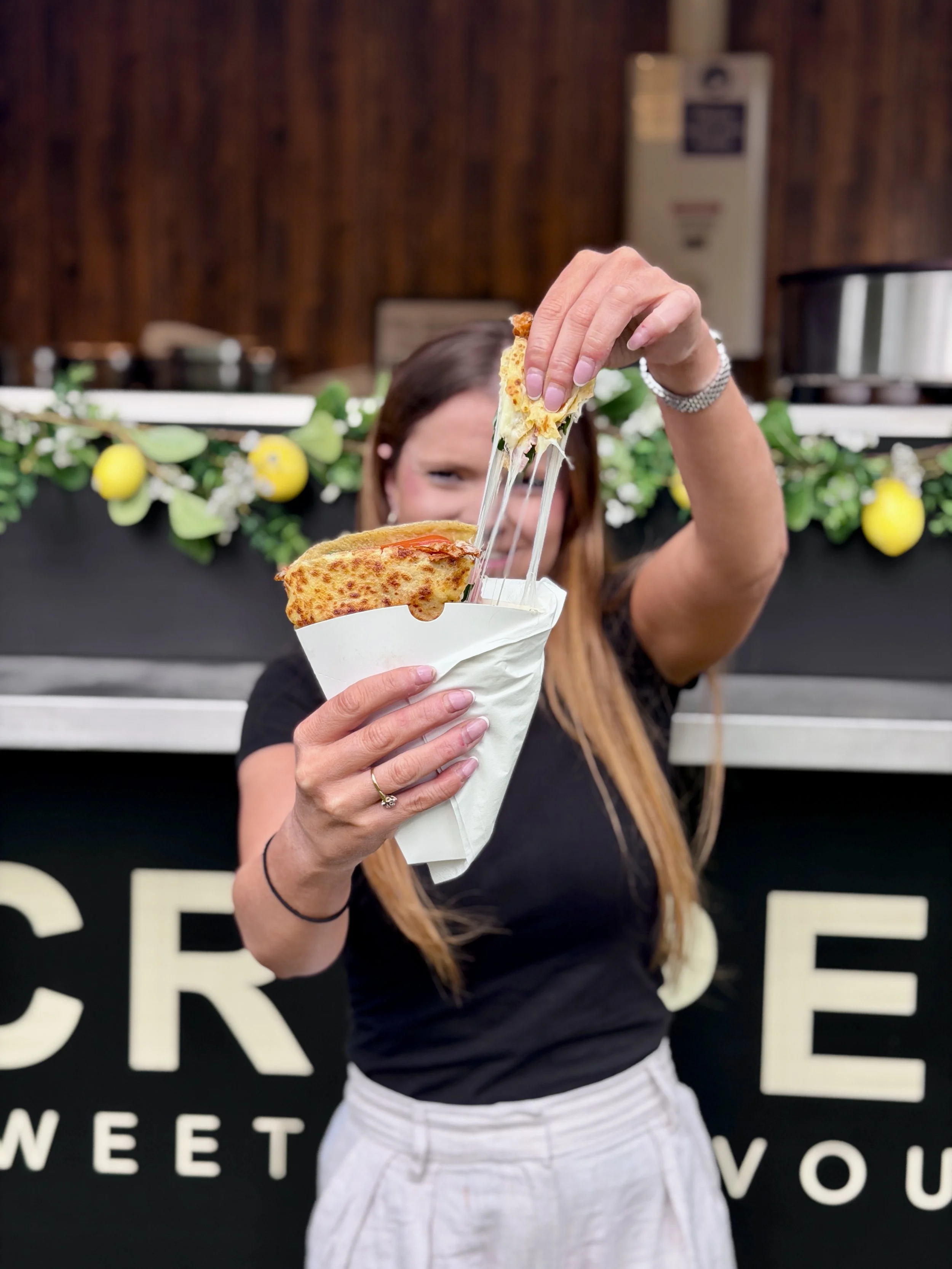 Person holding a cheese crepe with melted cheese stretching, standing in front of the crepe van with white lettering and decorations.