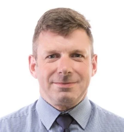 Headshot of a middle-aged man with short brown hair, wearing a blue checked shirt and a dark tie, smiling softly against a white background.