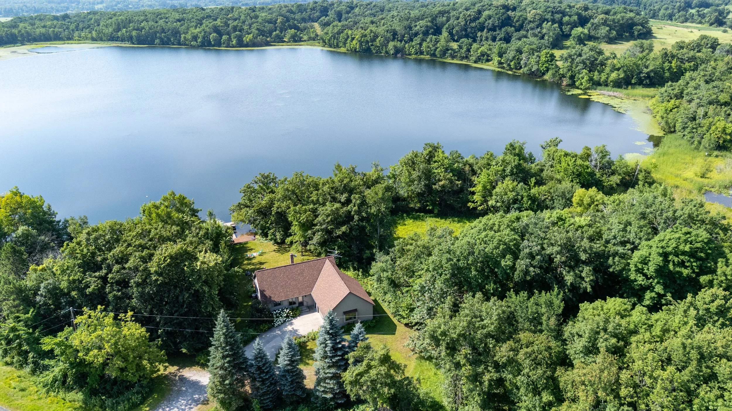 Aerial view of a lake surrounded by dense green trees, with a house and backyard at the bottom of the image.