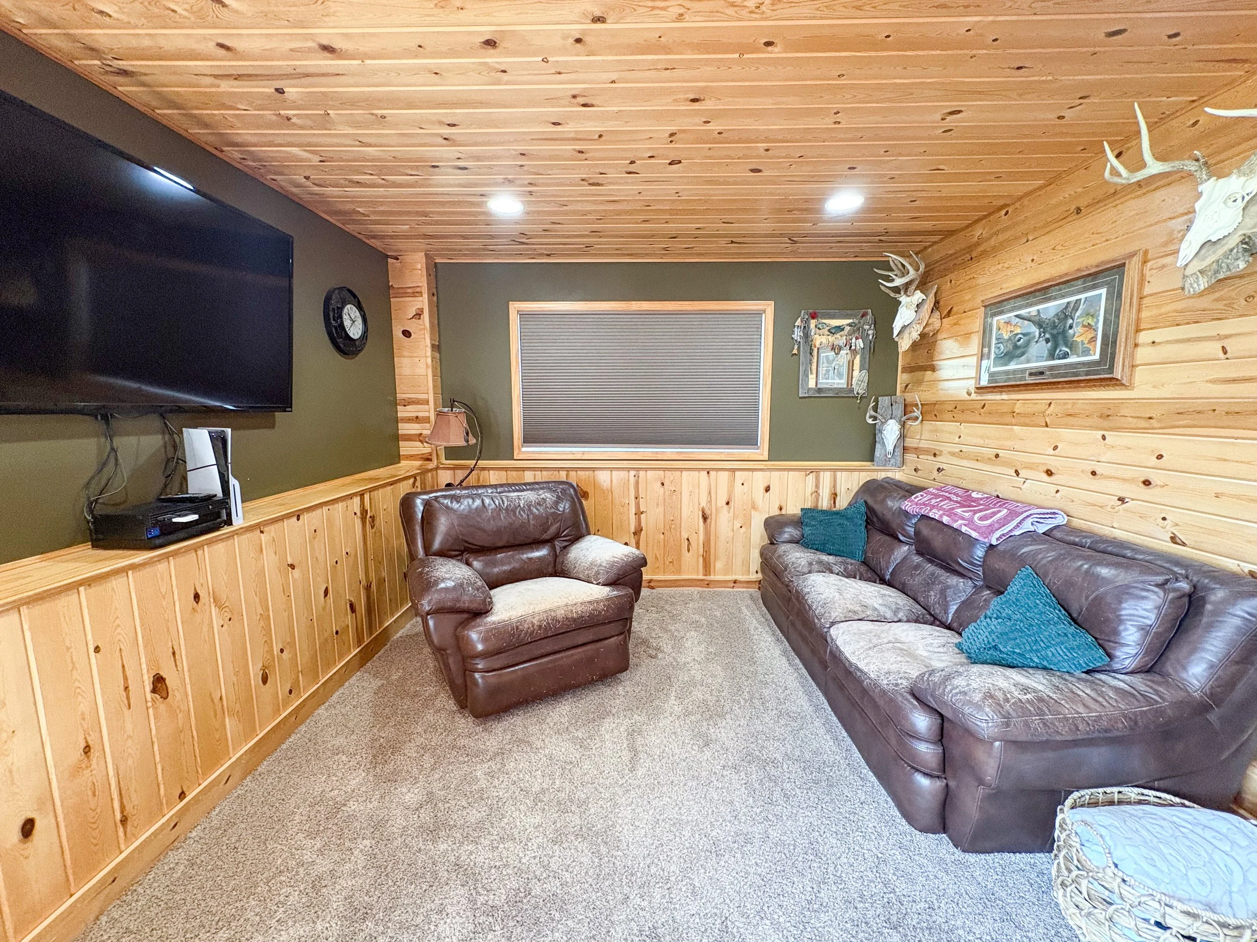 A cozy living room with wood-paneled walls and ceiling, a wall-mounted TV, a wall clock, a window with blinds, a small lamp, a leather armchair, a patterned tile table, and a sectional sofa with throw pillows and blanket, decorated with mounted deer 