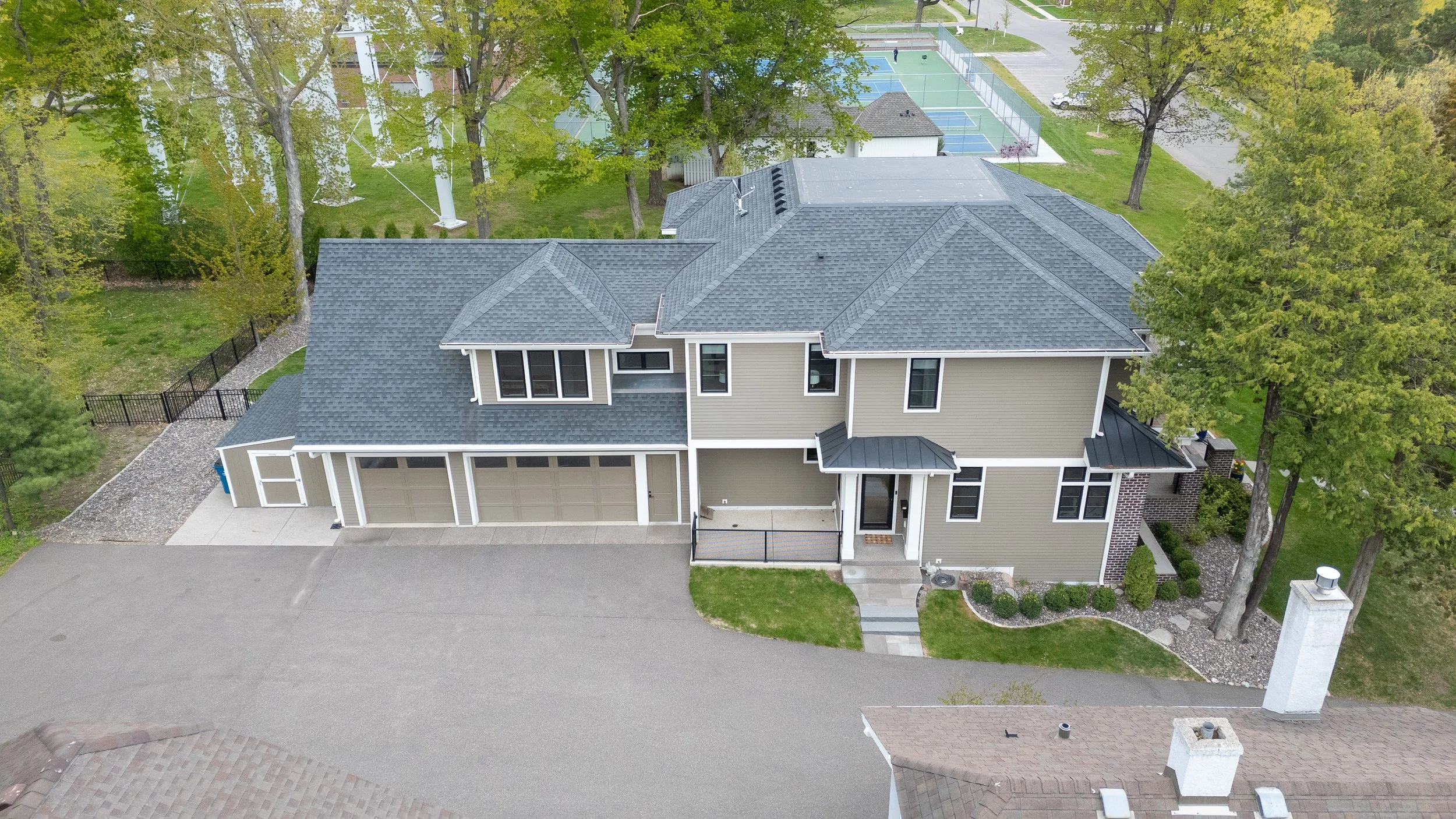Aerial view of a modern two-story house with a gray roof, beige exterior, and a double garage. The front yard includes a small lawn, a concrete pathway, and a black fence on one side. Surrounding the house are tall trees and neighboring properties wi