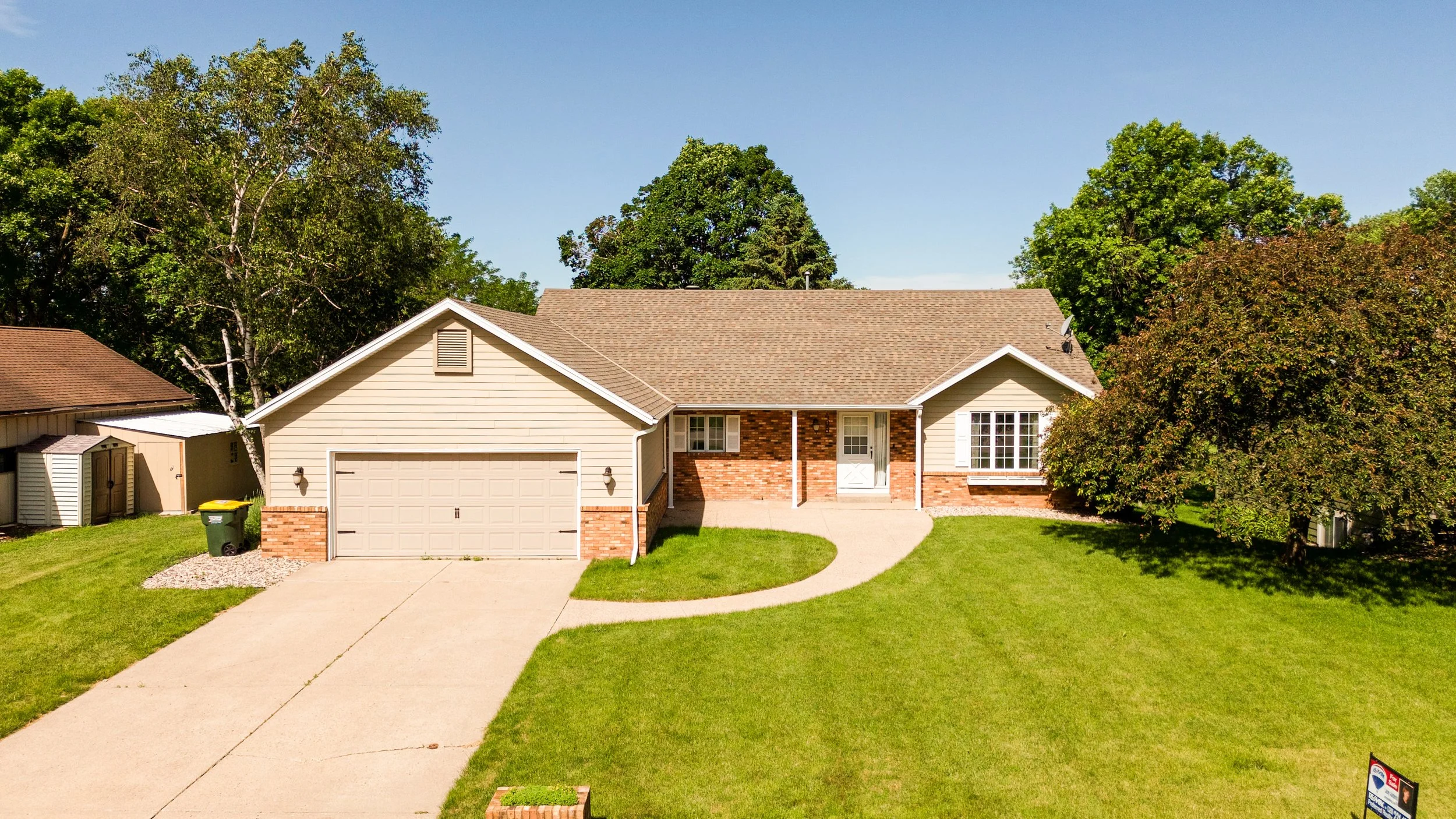 Front view of a suburban house with a beige garage door, brick accents, a small front porch, and surrounding green lawn and trees under a clear blue sky.