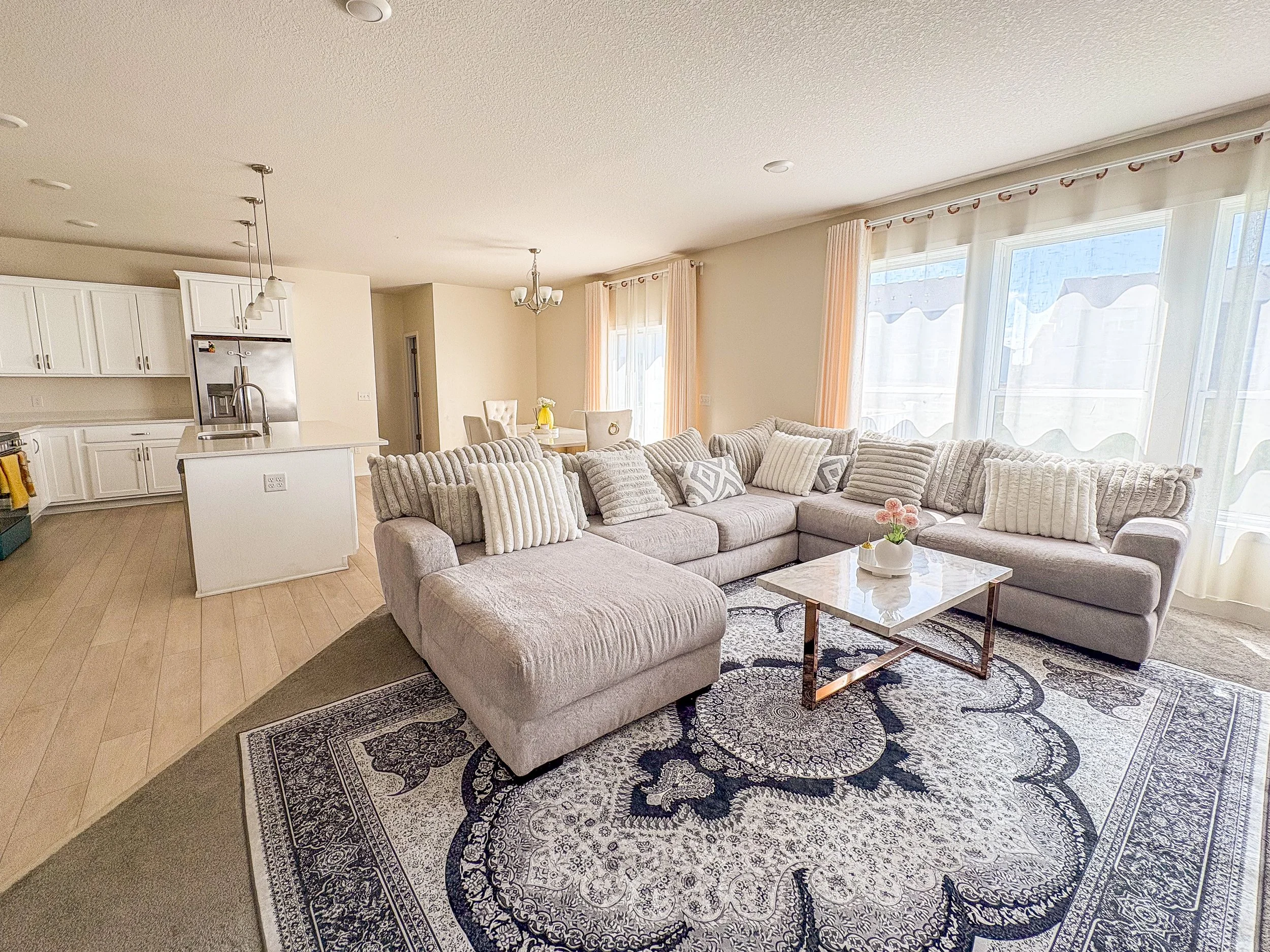 Bright living room with large windows, gray sectional sofa with patterned pillows, a black and white patterned rug, a glass coffee table, and a dining area with a chandelier and a vase of flowers. The kitchen with white cabinets and a stainless steel