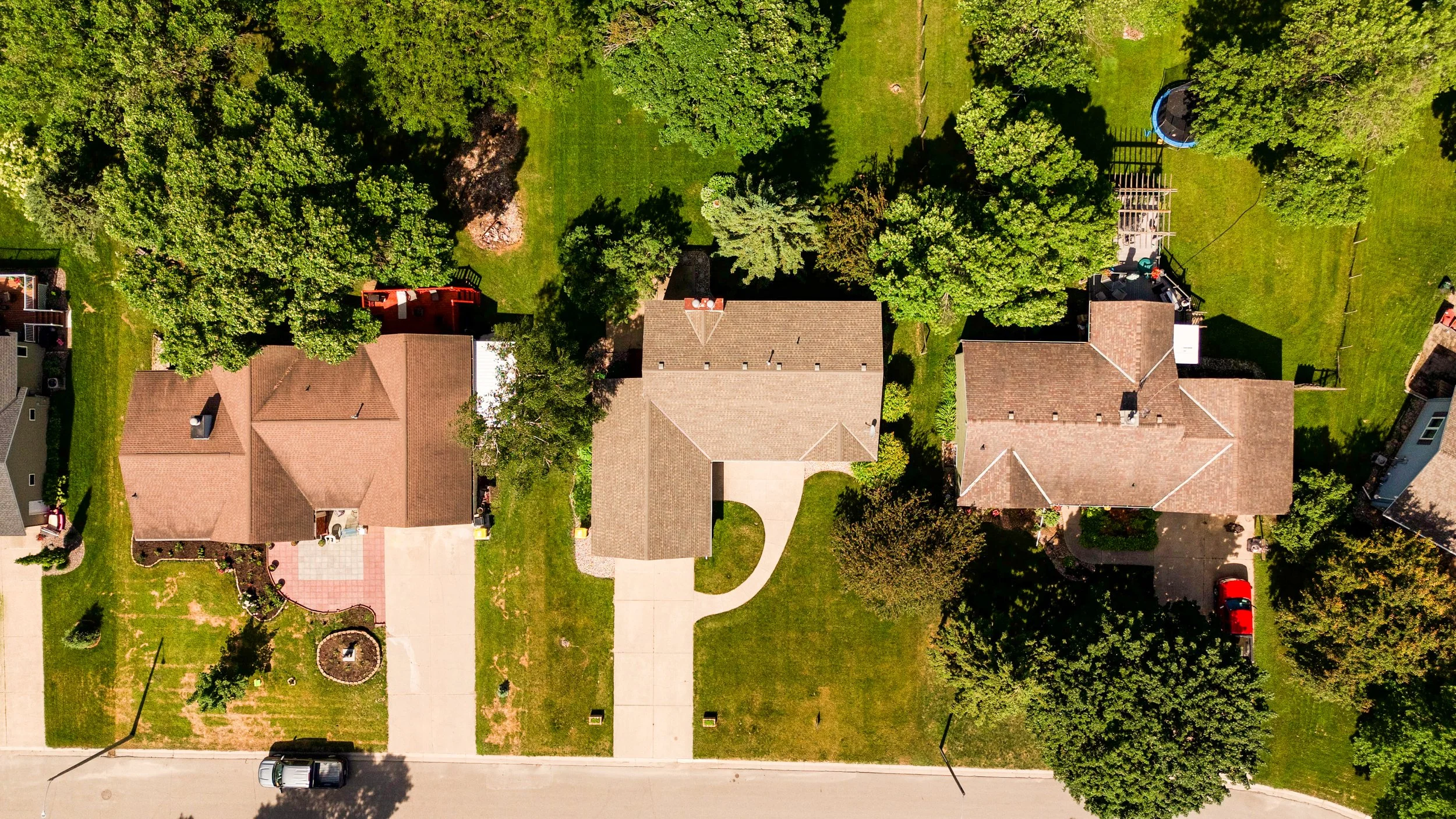 An aerial view of a suburban neighborhood showing houses with tiled roofs, driveways, green lawns, trees, and a backyard with a trampoline and a wooden deck.