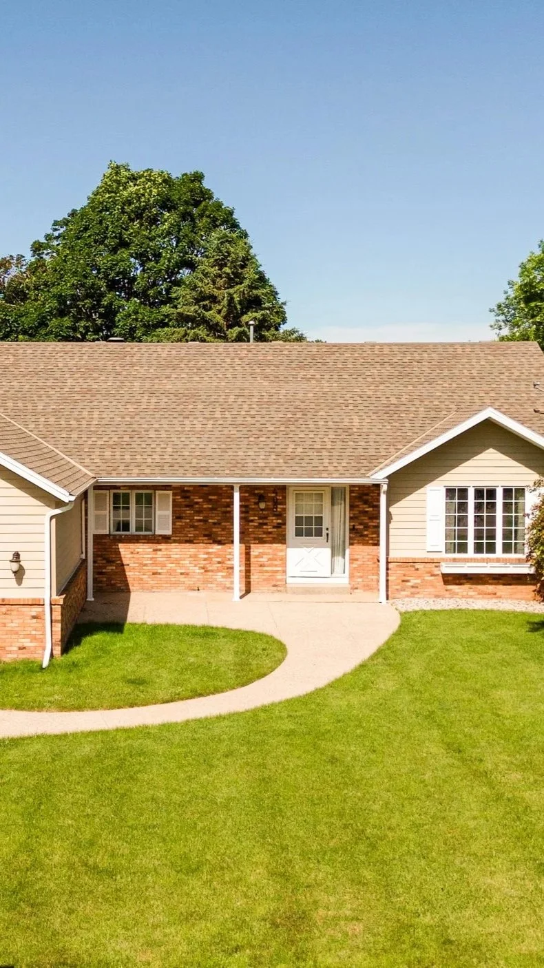 Front view of a single-story house with a brick and beige siding exterior, white door, large windows, manicured lawn, and a curved concrete walkway leading to the entrance. Tall trees and a clear blue sky in the background.