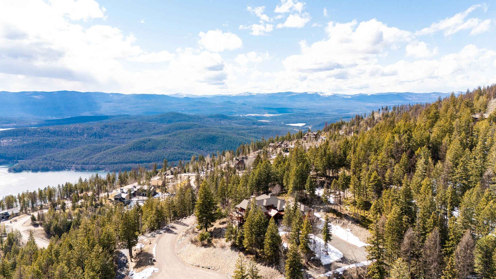 A scenic mountain landscape with forested hills, snow patches, and a winding road with houses, under a partly cloudy sky.