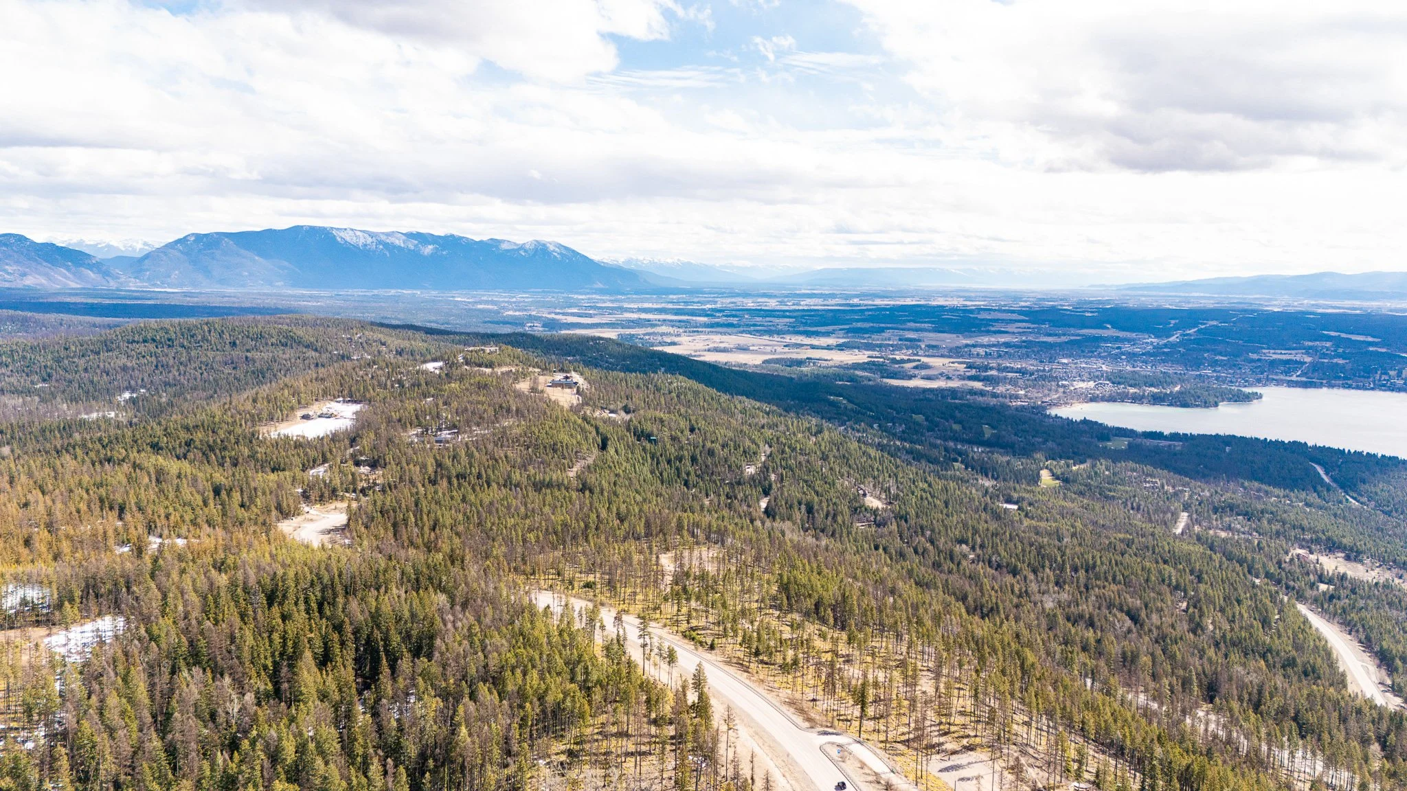 An aerial view of a forested landscape with mountains in the background, a lake, and a winding road.
