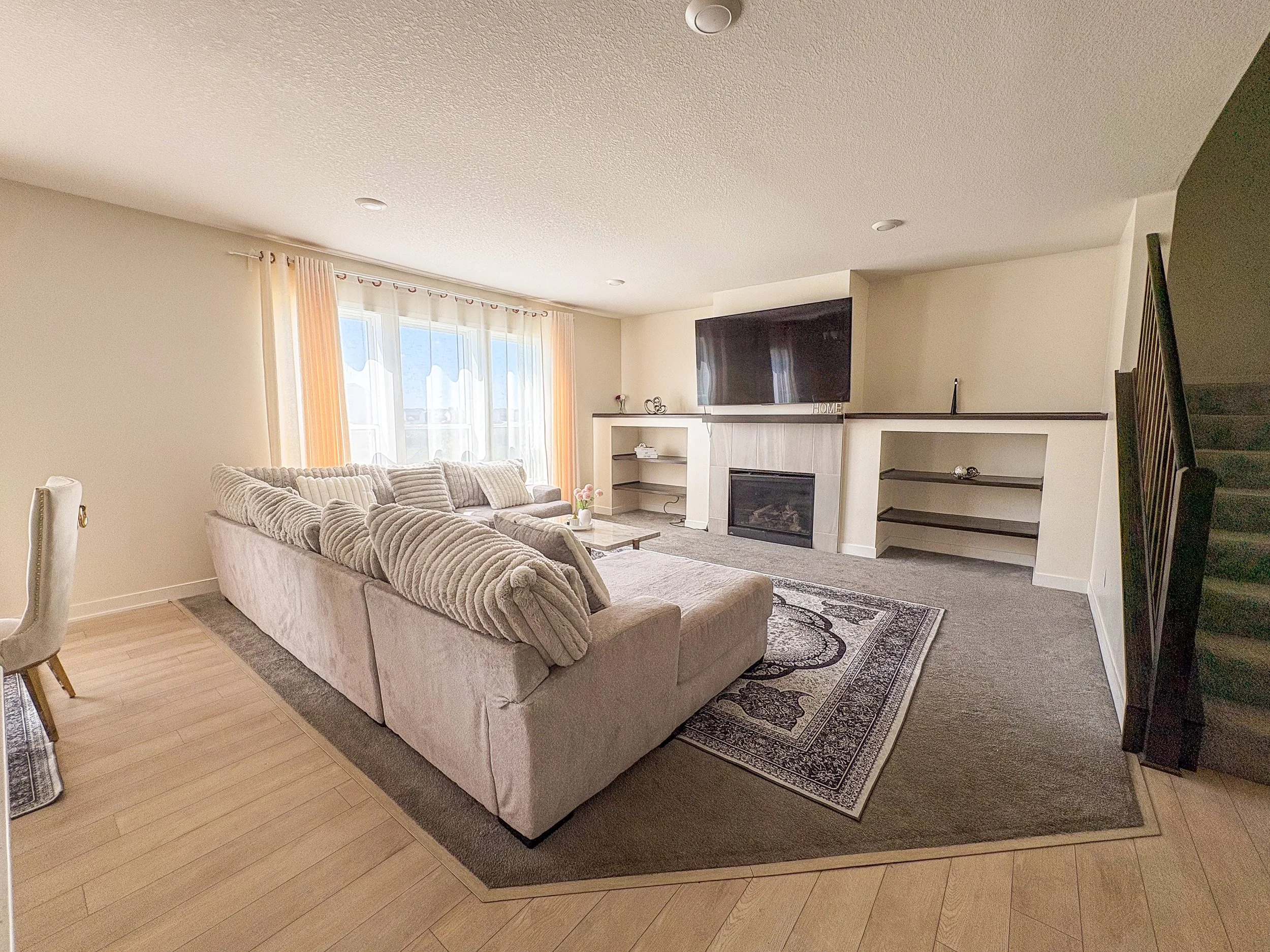 Living room with beige sofa, fireplace, TV on the wall, large window with curtains, staircase to the right, with decorative items on the mantle and shelves.