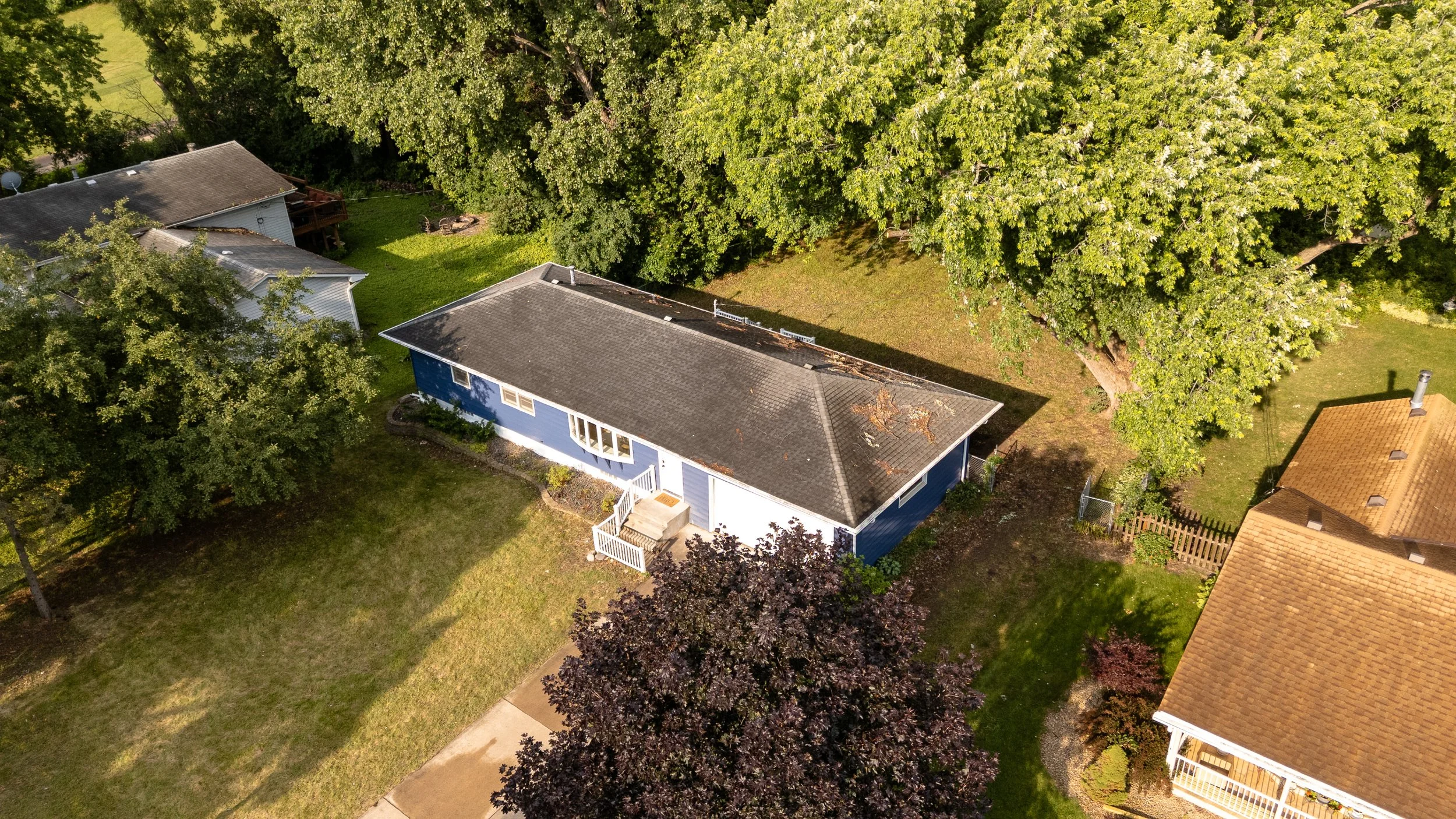 An aerial view of a house with a gray roof, surrounded by large green trees and a lawn, in a suburban neighborhood during daytime.