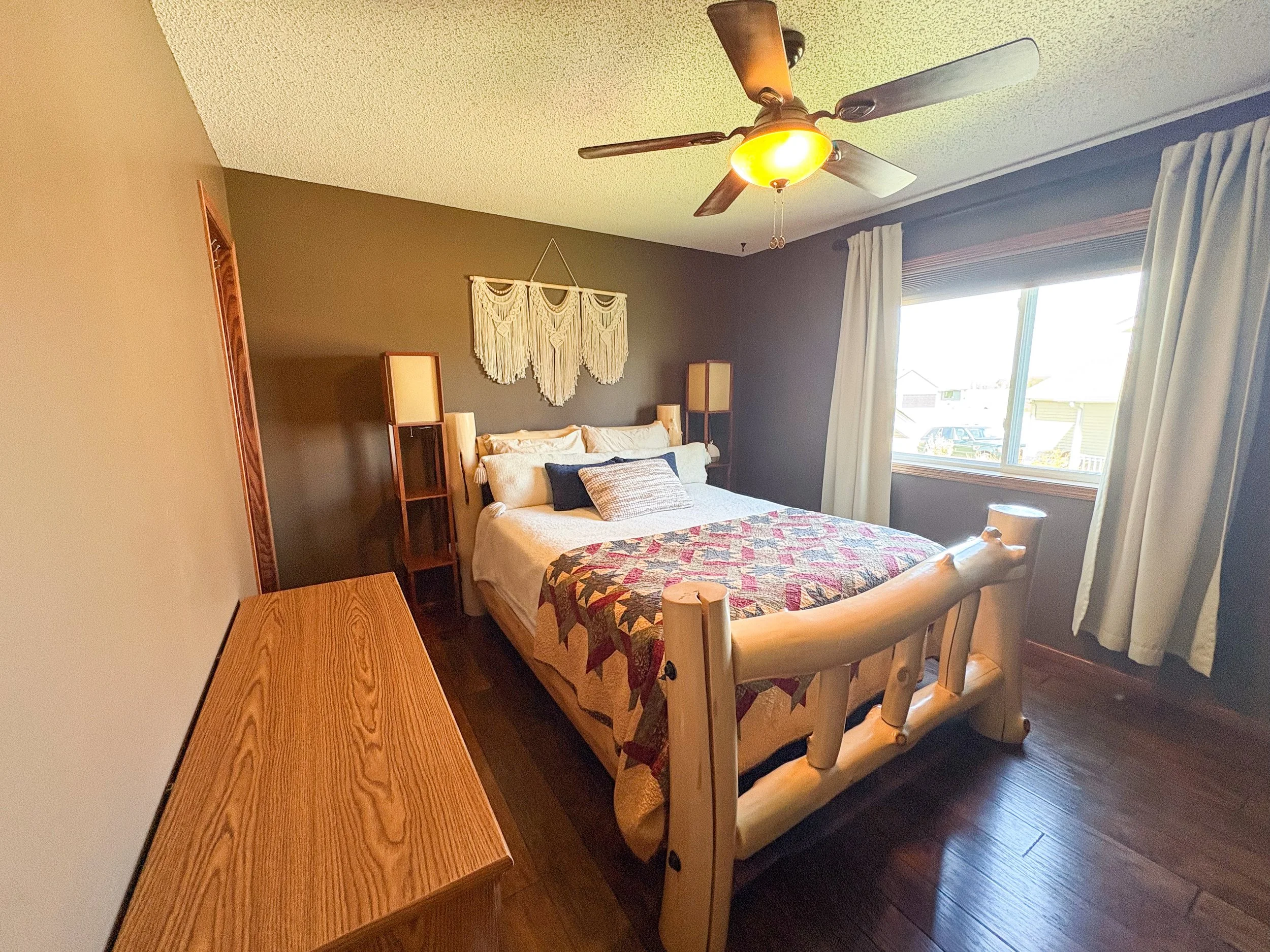 A cozy bedroom featuring a wooden bed with a quilted bedspread, pillows, and a nightstand with a lamp. There are two wooden shelves on each side of the bed and a macrame wall hanging above it. The room has a window with white curtains and a ceiling f