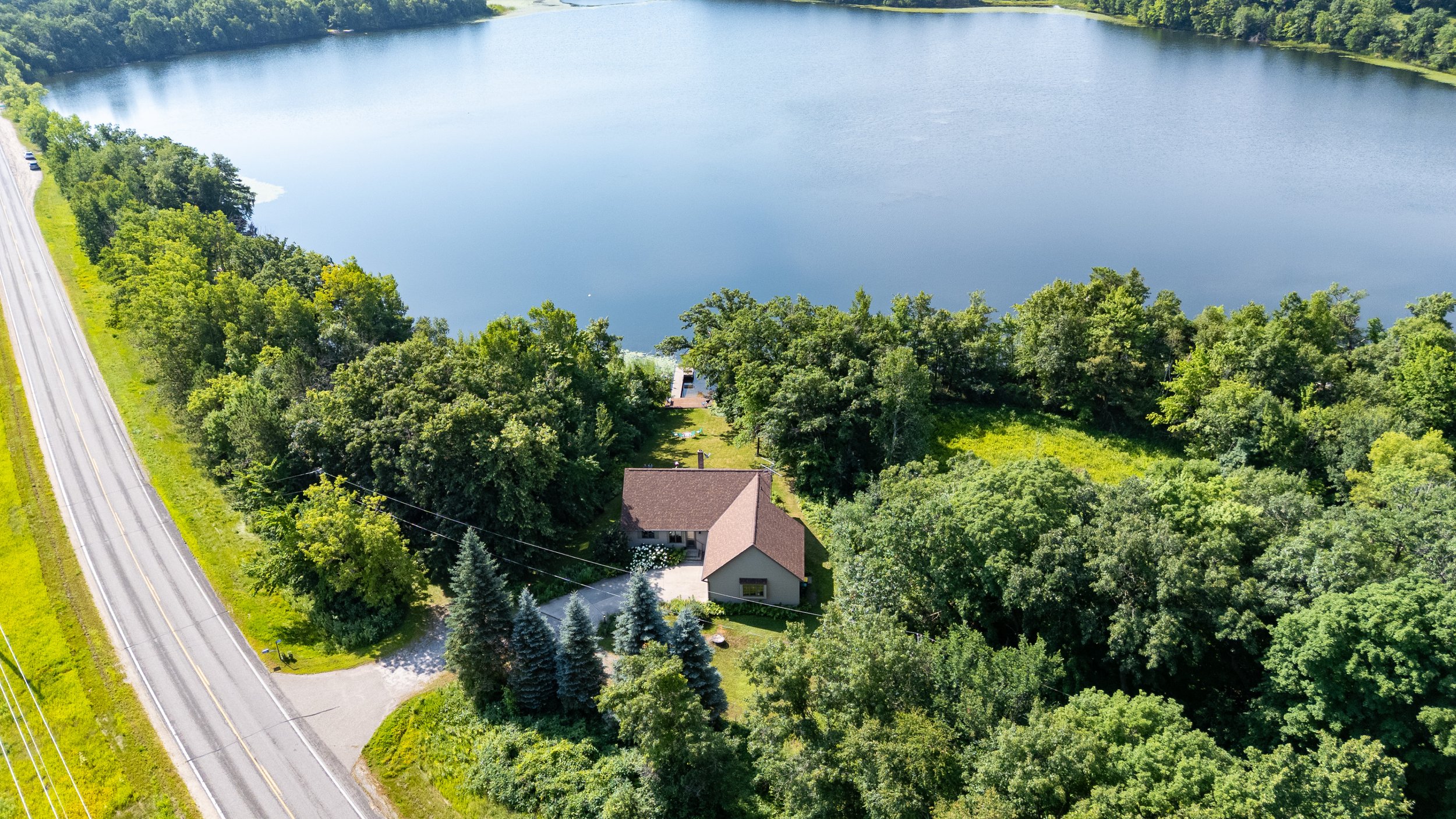 Aerial view of a house surrounded by trees near a large lake and a road with a few parked cars.