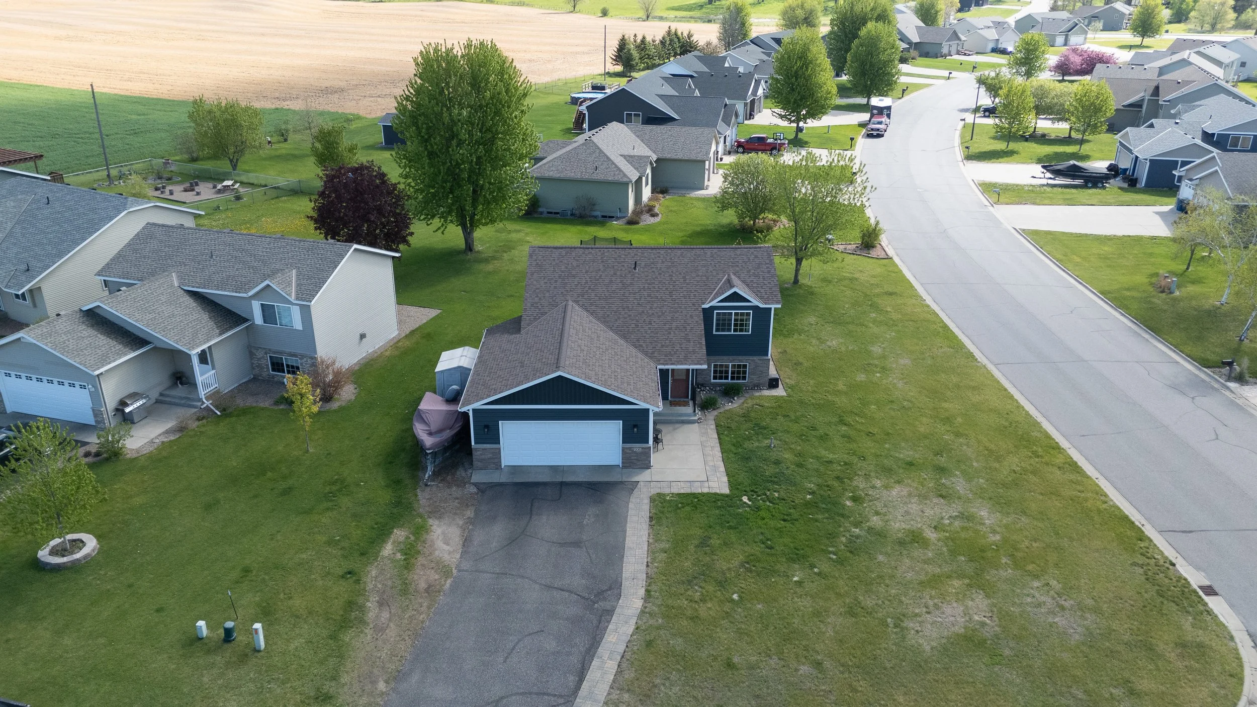 Aerial view of a suburban neighborhood with houses, green lawns, trees, and a curved street. One house in the foreground has a dark blue exterior, gray roof, and attached garage. The neighborhood is bordered by farmland.